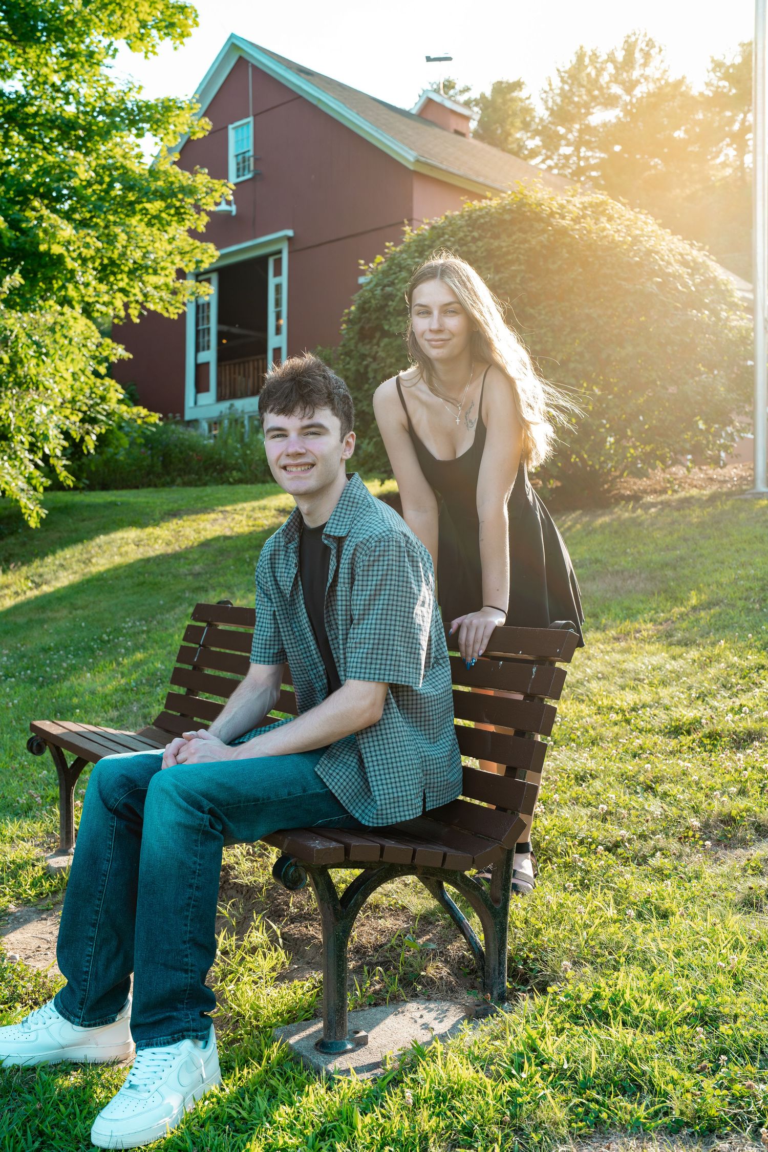 Teen boy and girl posing on a wooden bench in a sunny backyard with a red house backdrop.