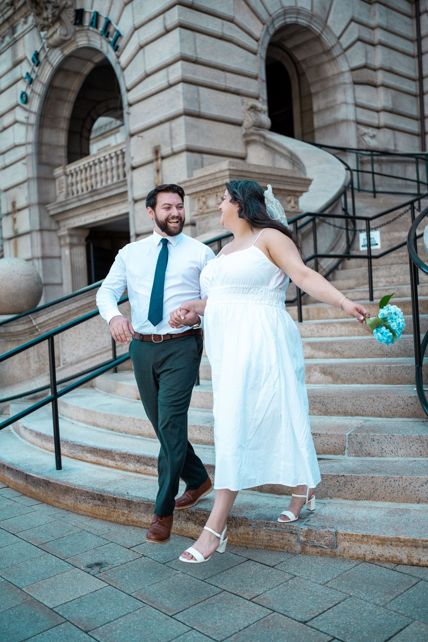 Bride and groom walk down stone steps of historic Worcester City Hall, holding hands and smiling, bride holding blue bouquet. Elopment.
