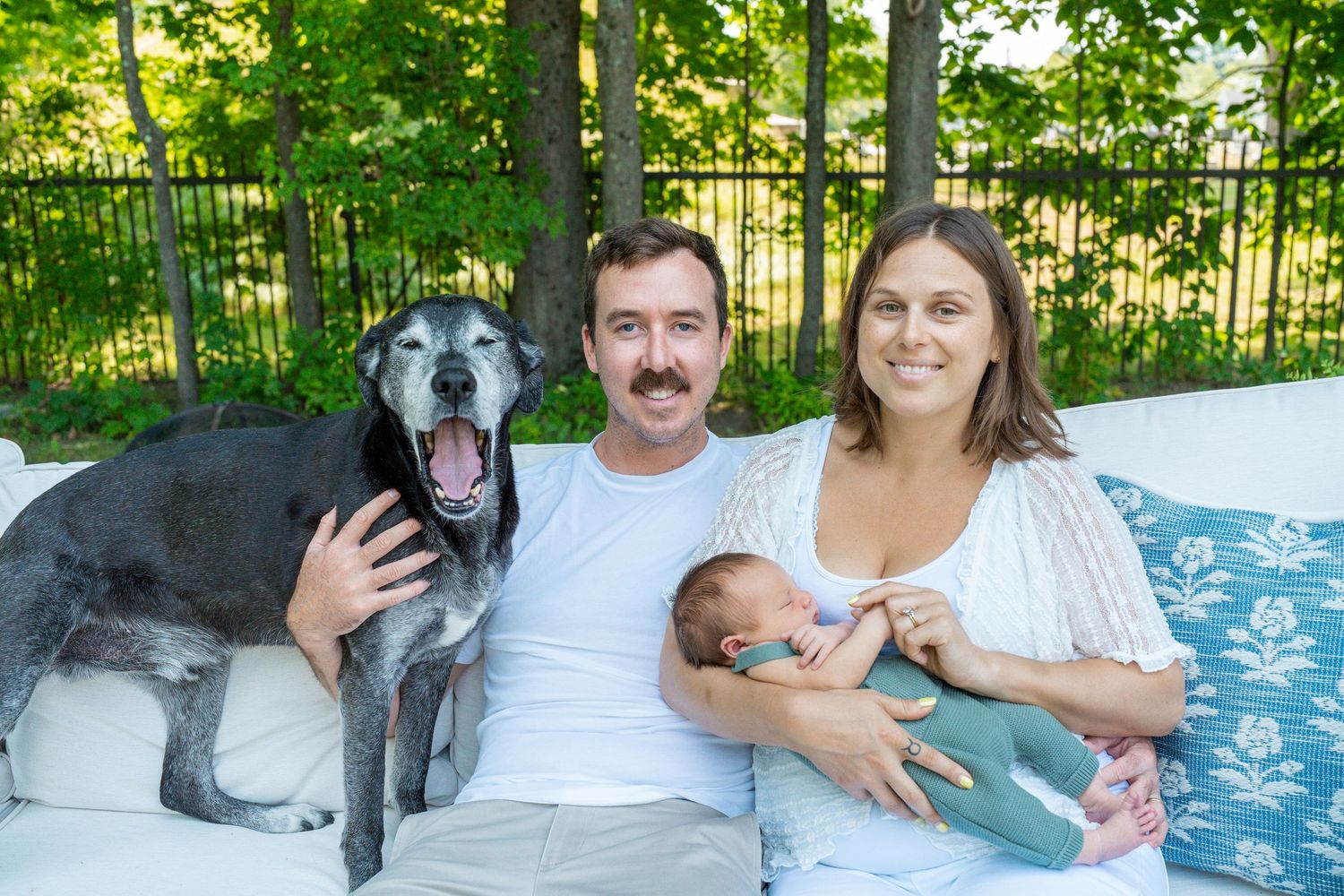 Family portrait with couple, newborn baby, and senior gray dog sitting on outdoor white sofa with green trees.