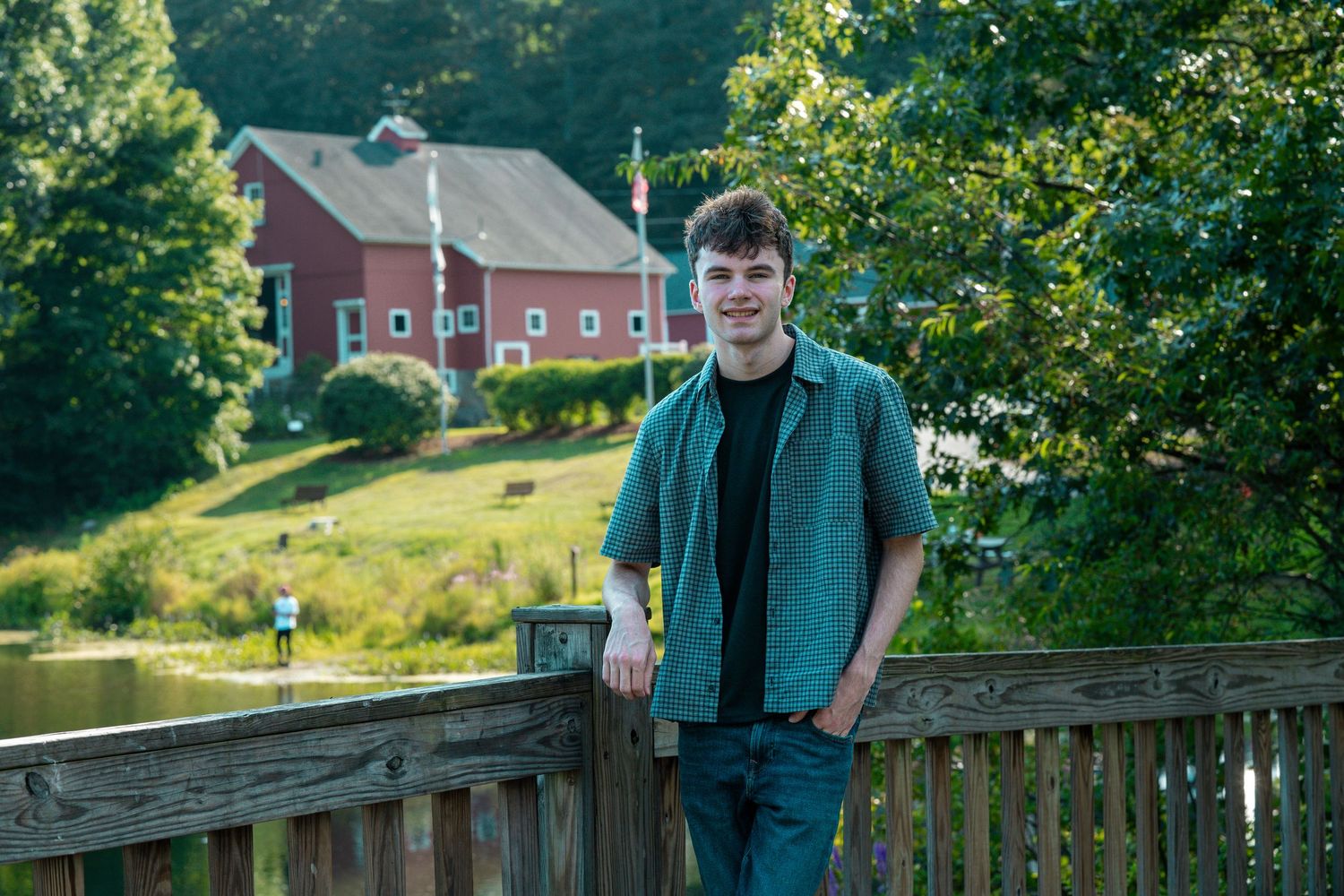Young man smiling on a wooden deck with a red barn and lush green trees in the background.
