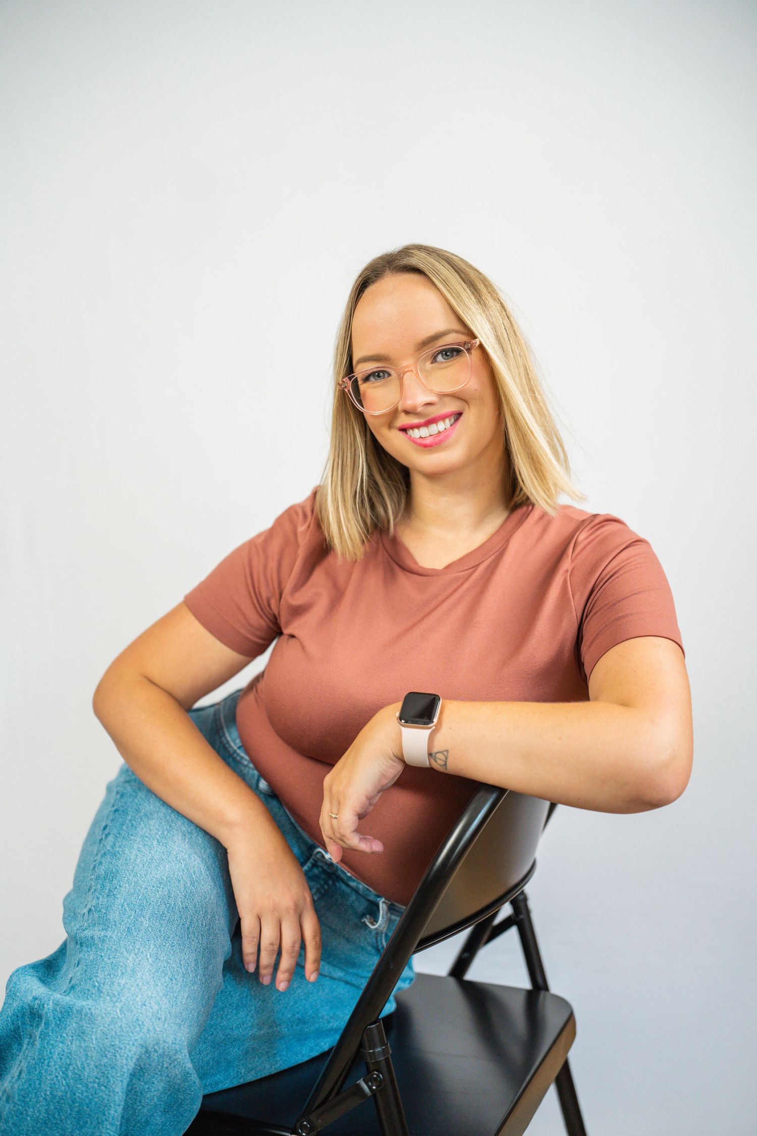 Smiling blonde woman in glasses, pink t-shirt, and jeans sitting on a chair with an Apple Watch on her wrist.