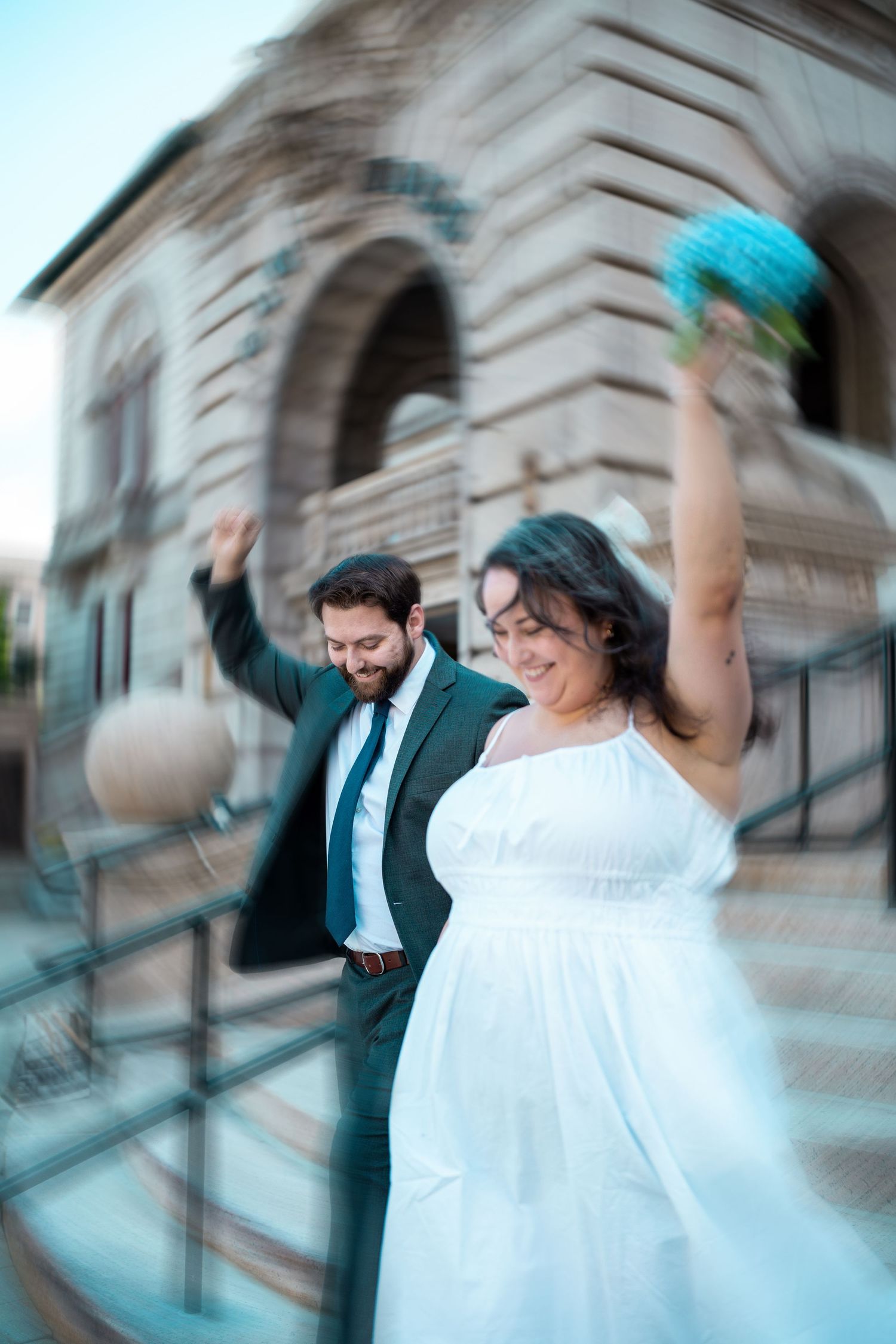 Joyful bride white dress raises bouquet while groom in dark suit celebrates on steps of elegant stone building. Elopement in Worcester, MA