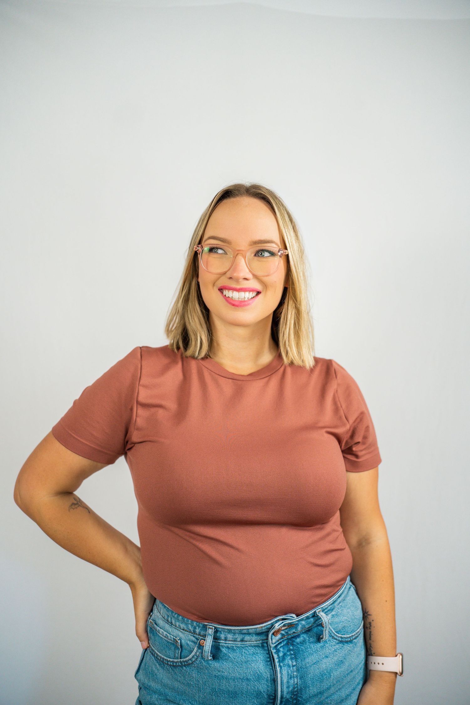 Smiling blonde woman wearing glasses, a rust-colored t-shirt, and jeans, posing confidently against a white background.