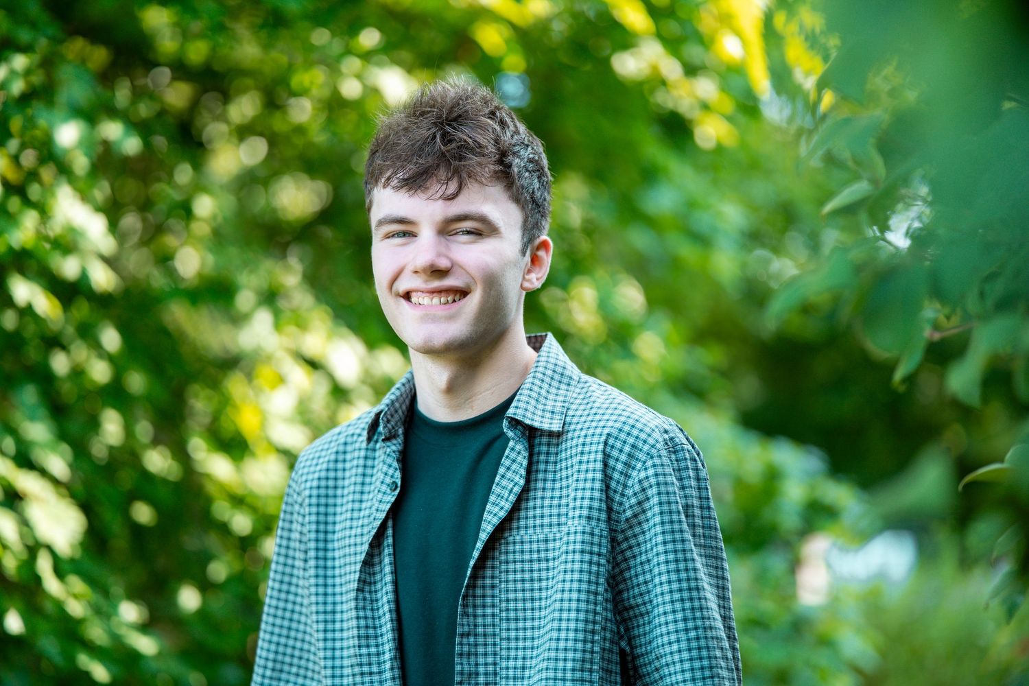 Smiling young man in plaid shirt standing outdoors among lush green trees and bokeh foliage background.