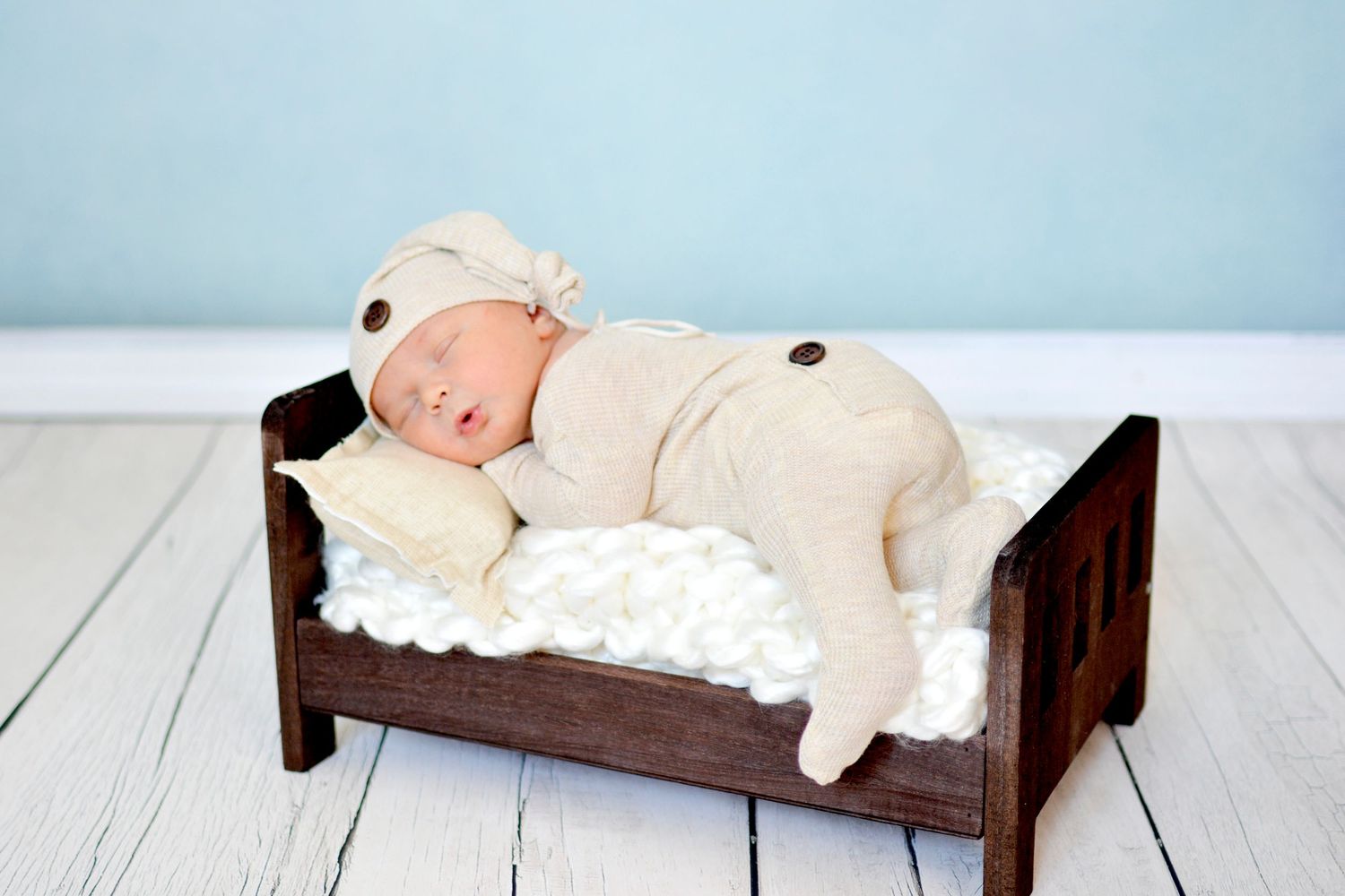 Newborn baby sleeping peacefully in a tiny wooden bed, wearing a cream bear outfit and hat.