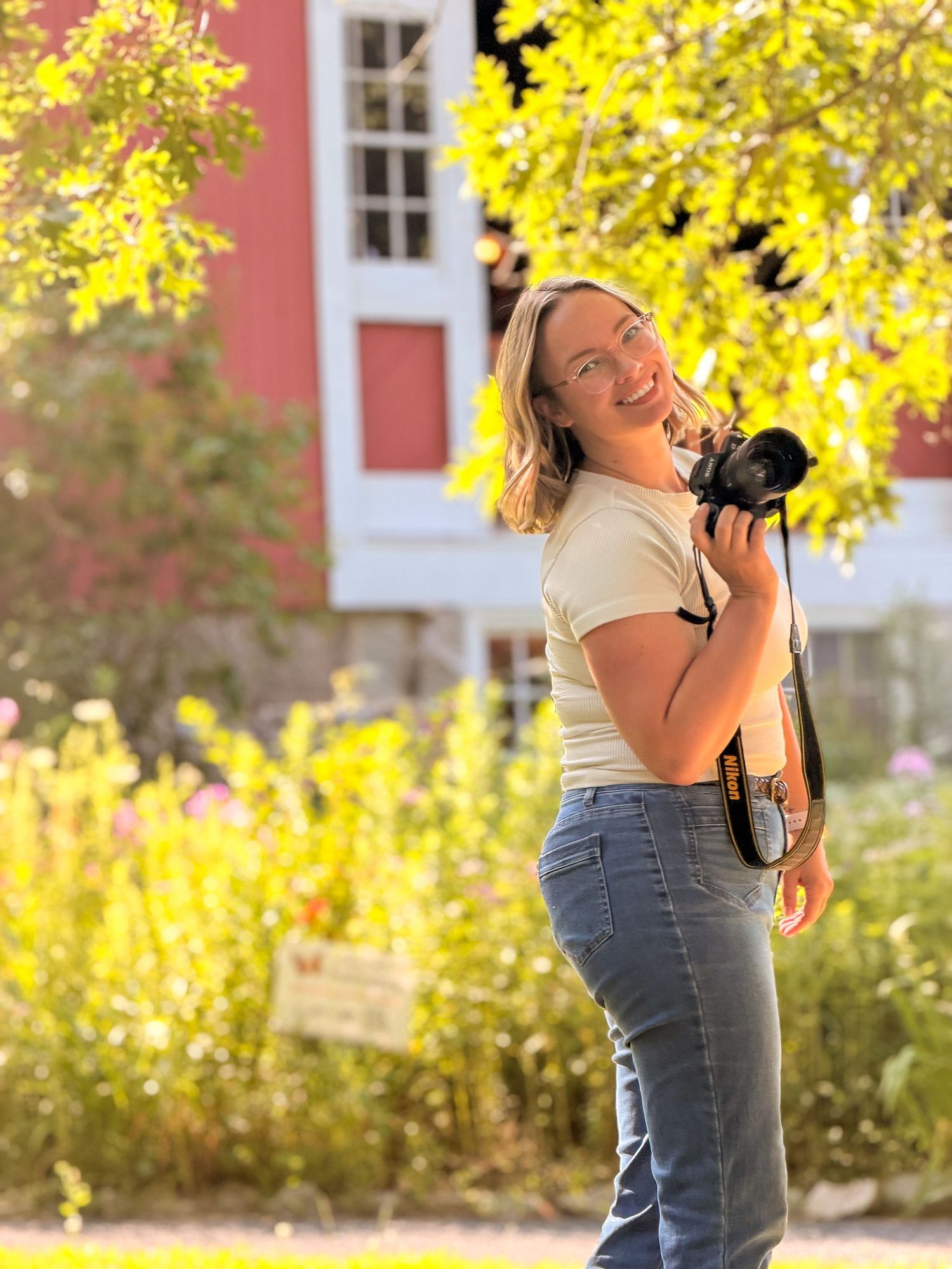 Smiling blonde woman holding a camera outdoors near colorful flowers and a red building on a sunny day. Catrina Gardner at Studio Joki.