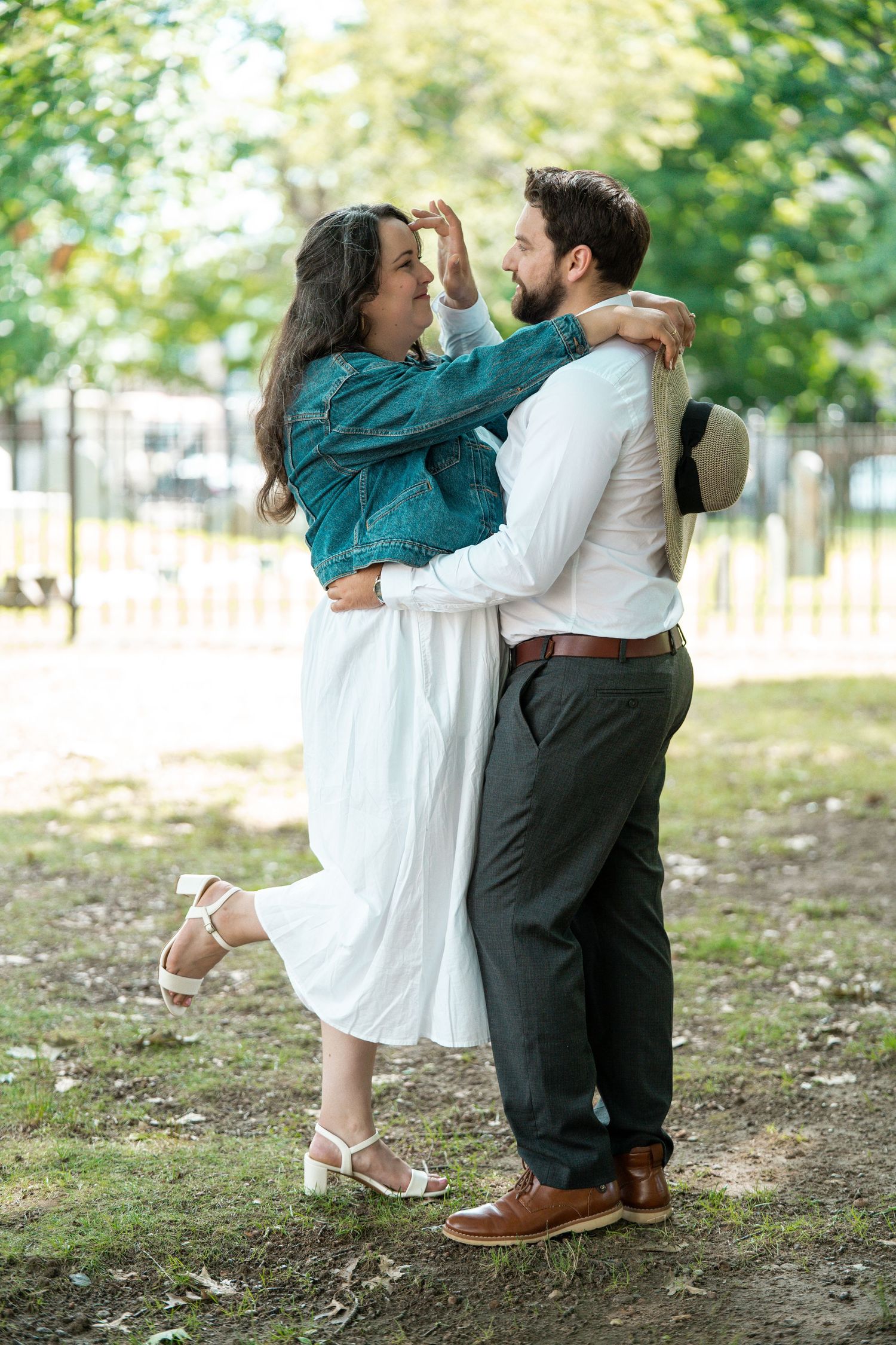 A couple embraces and dances together in a lush green park, woman in white dress and denim jacket. Worcester City Hall.
