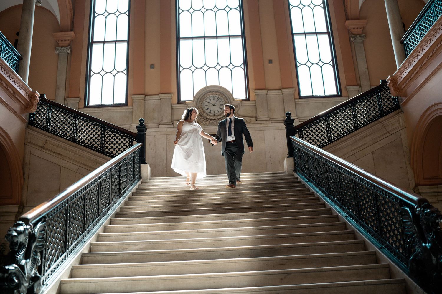 Bride and groom walking up grand staircase in elegant building with ornate railings and tall windows. Elopement at Worcester City Hall.