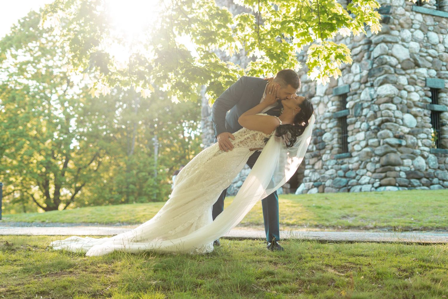 Couple sharing romantic dip kiss in wedding attire on green lawn with stone castle and sunlit trees.