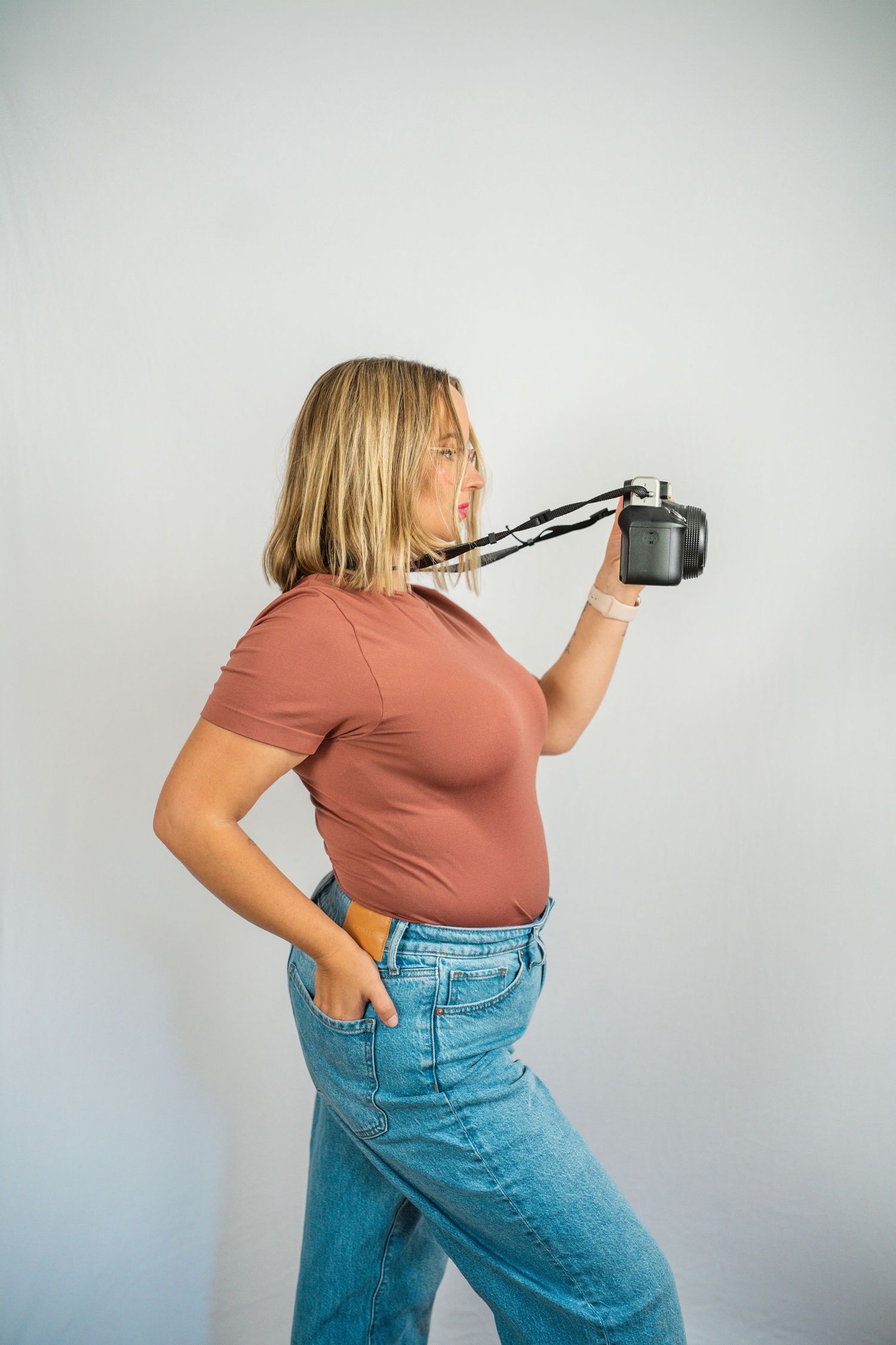 Blonde woman in brown t-shirt and jeans holding a camera with strap against white background.