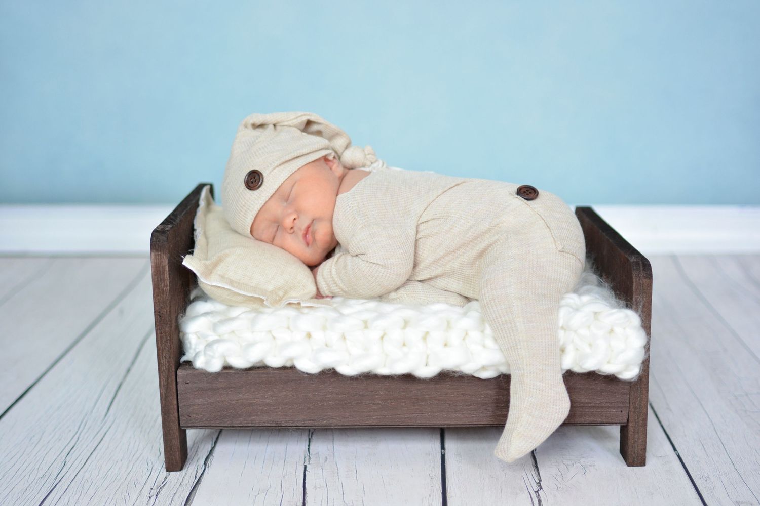 Newborn baby sleeping peacefully in a tiny wooden bed wearing a beige knit outfit and hat.