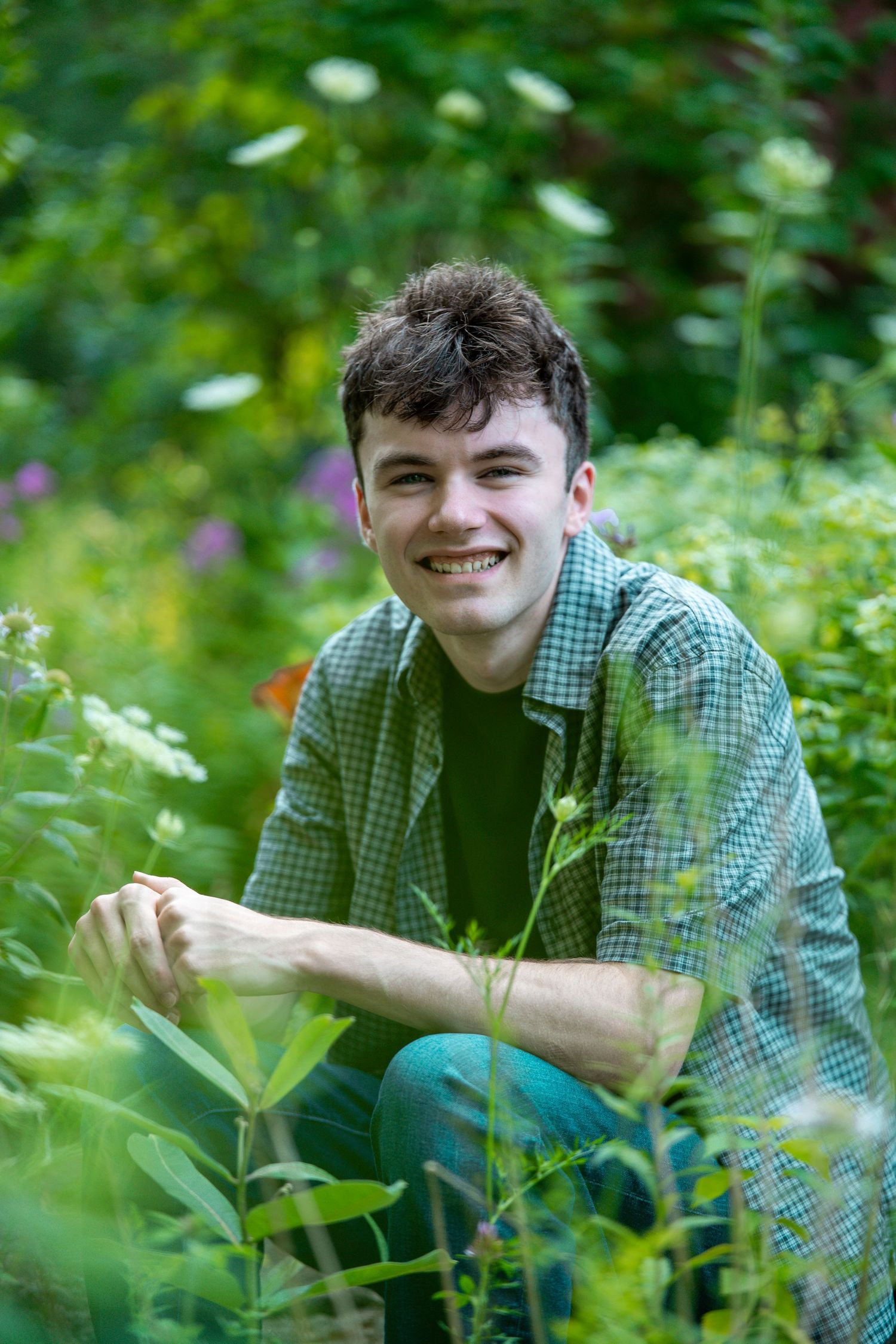 Young man smiling in a lush green garden, crouching among plants wearing a green plaid shirt.