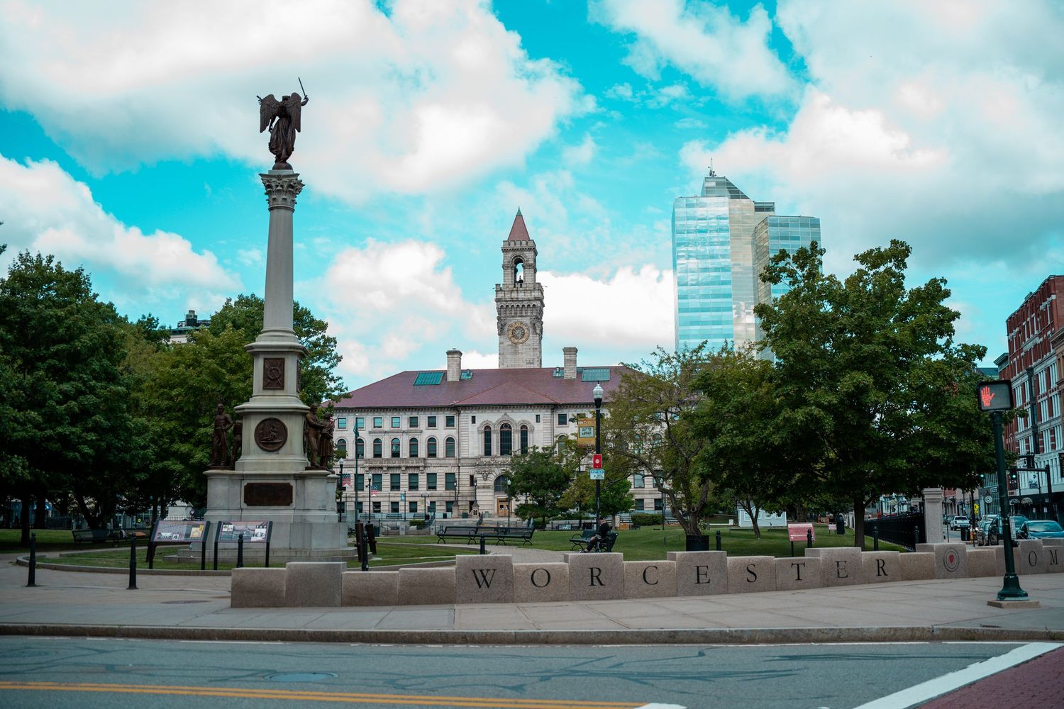 Worcester city square featuring a tall monument, historic city hall, and lush green trees under a cloudy sky. Massachusetts