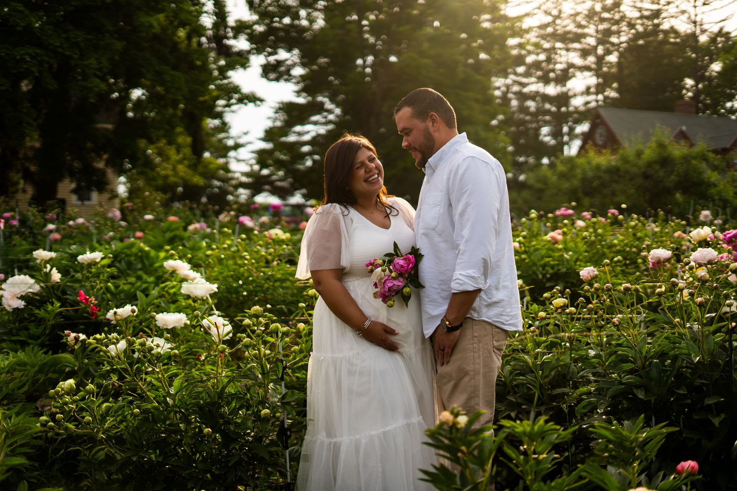 Couple in white attire smiling at each other in a lush garden with pink and white peonies in golden hour light.