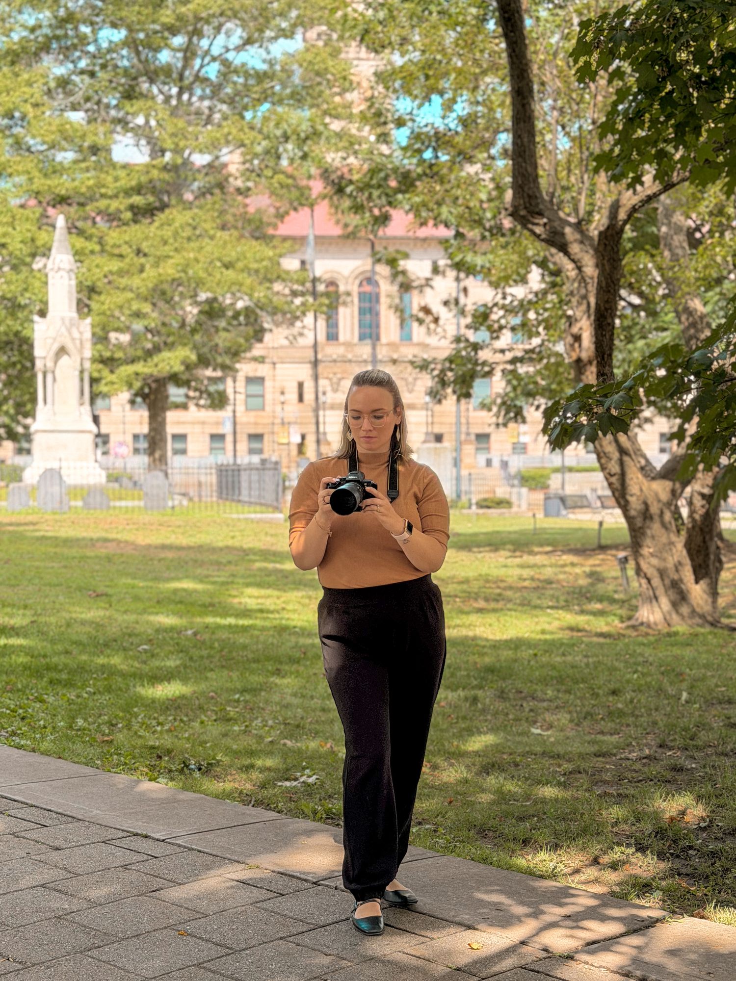 A young woman with braided hair holds a camera in a sunny park, with a historic building and monument in the background. Catrina Gardner.