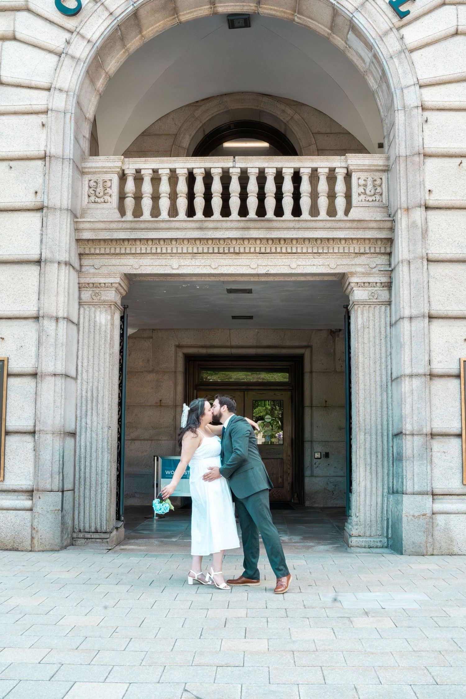 Bride and groom share a kiss under a grand stone archway with balustrade balcony at their wedding venue. Elopement at Worcester City Hall.