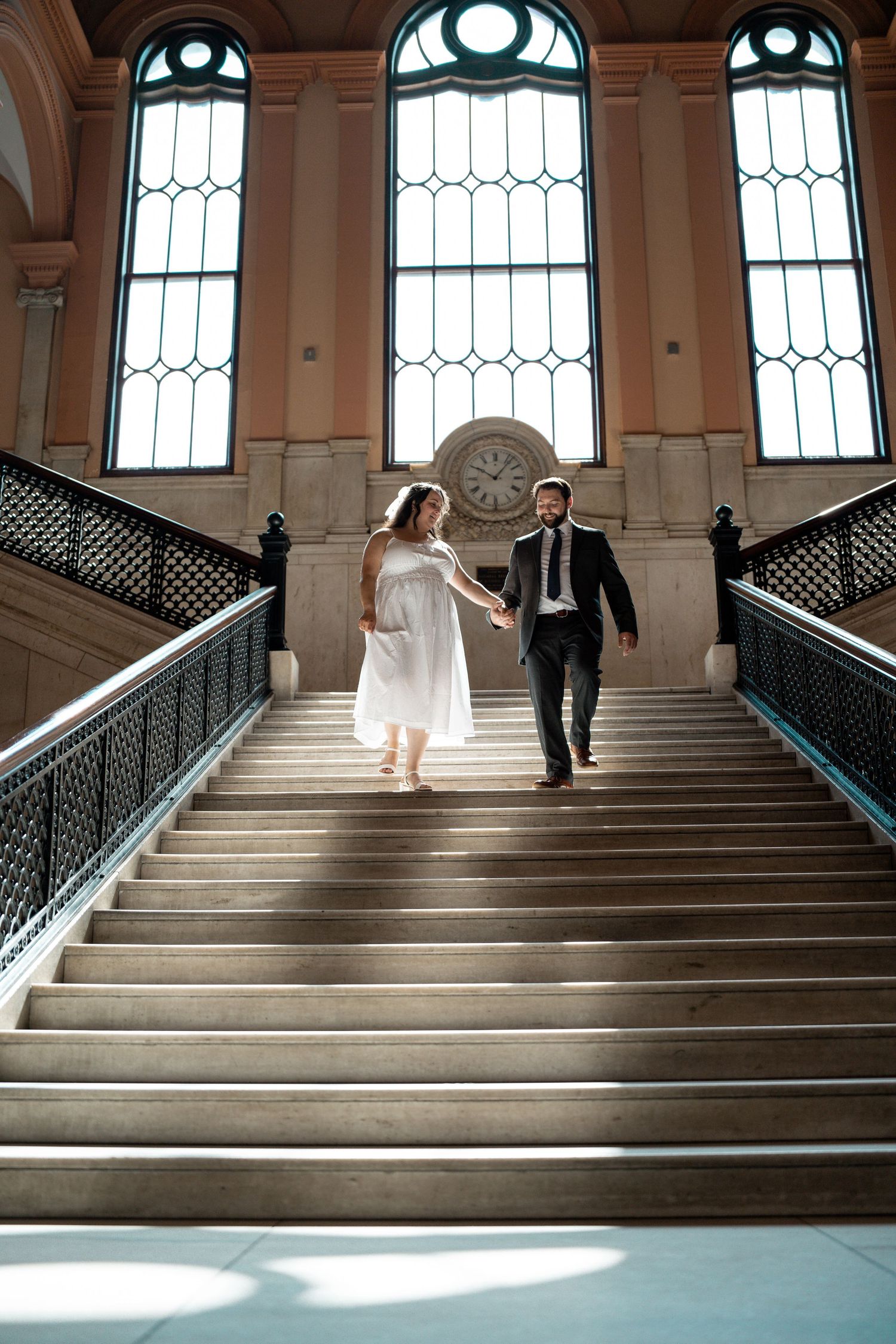 Bride and groom holding hands walking down grand staircase in elegant venue with arched windows and clock. Elopement at Worcester City Hall.