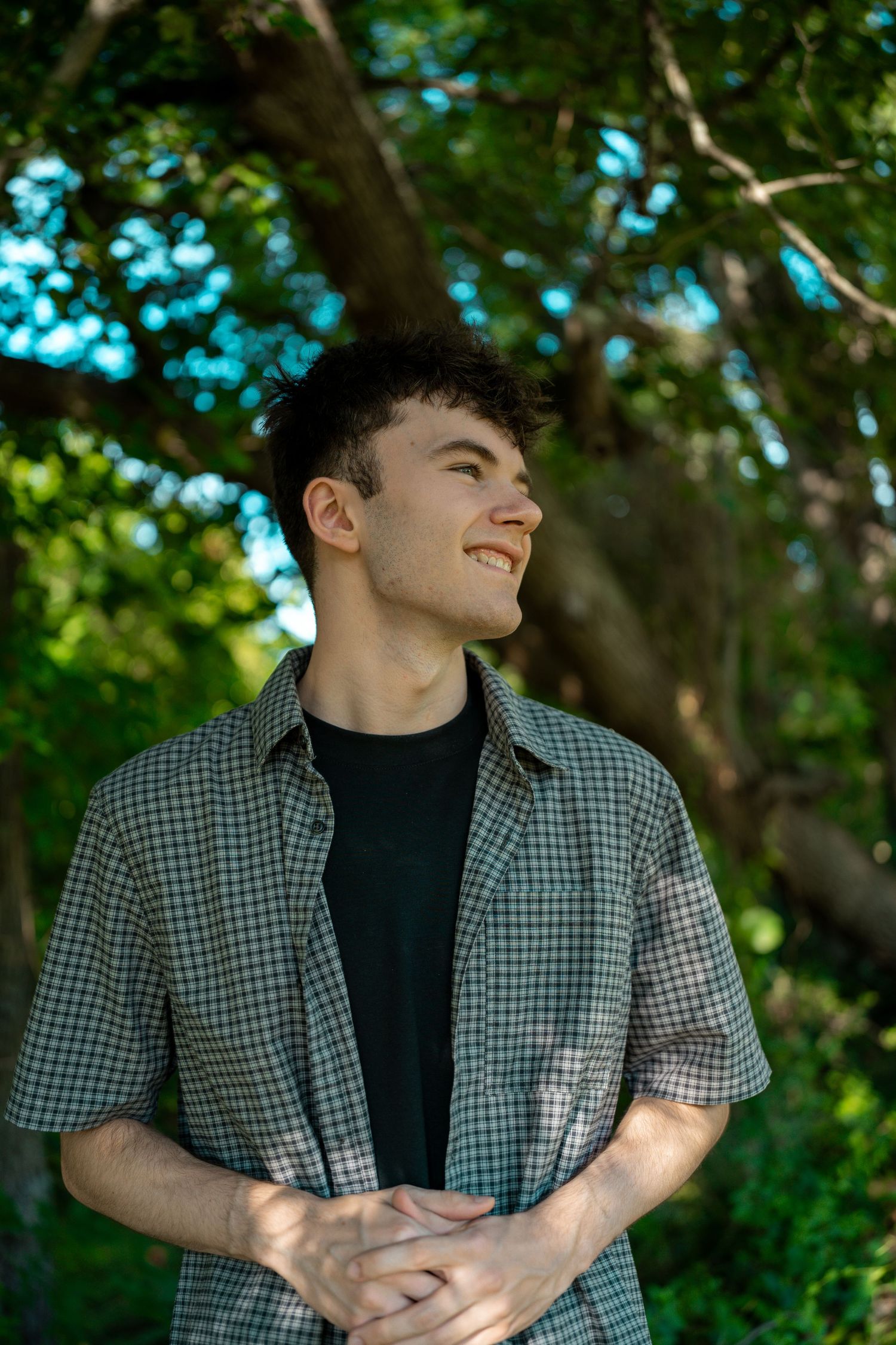Smiling young man with curly hair wearing a plaid shirt standing outdoors surrounded by lush green trees.