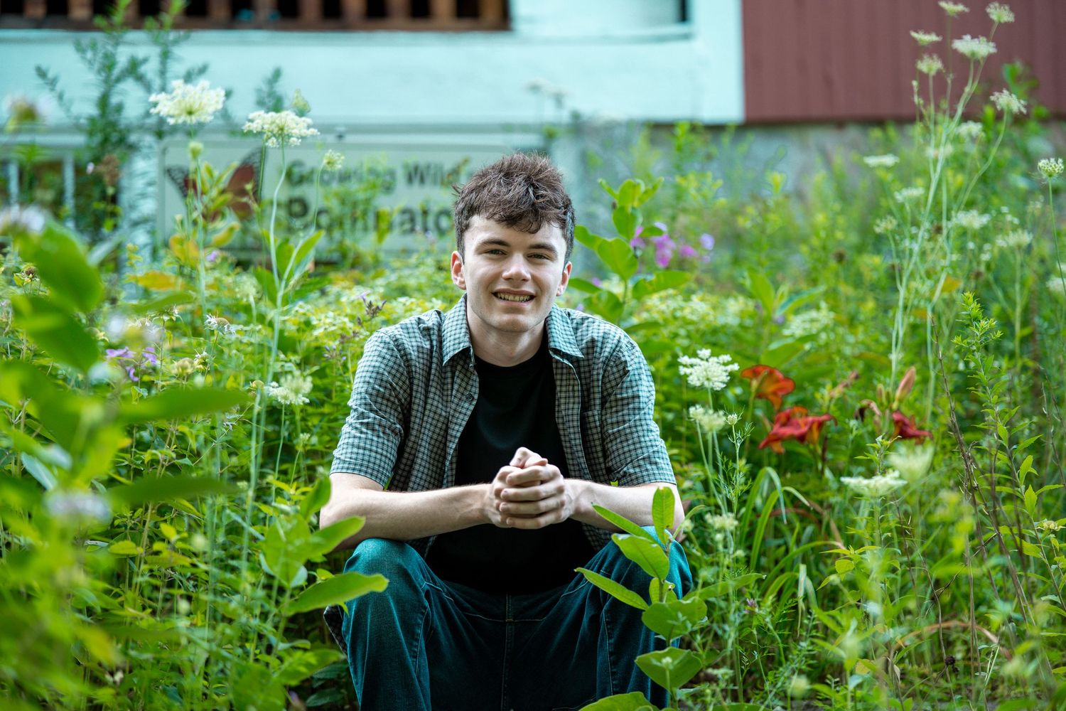Young man crouching in a lush garden surrounded by wildflowers and pollinator plants outdoors.