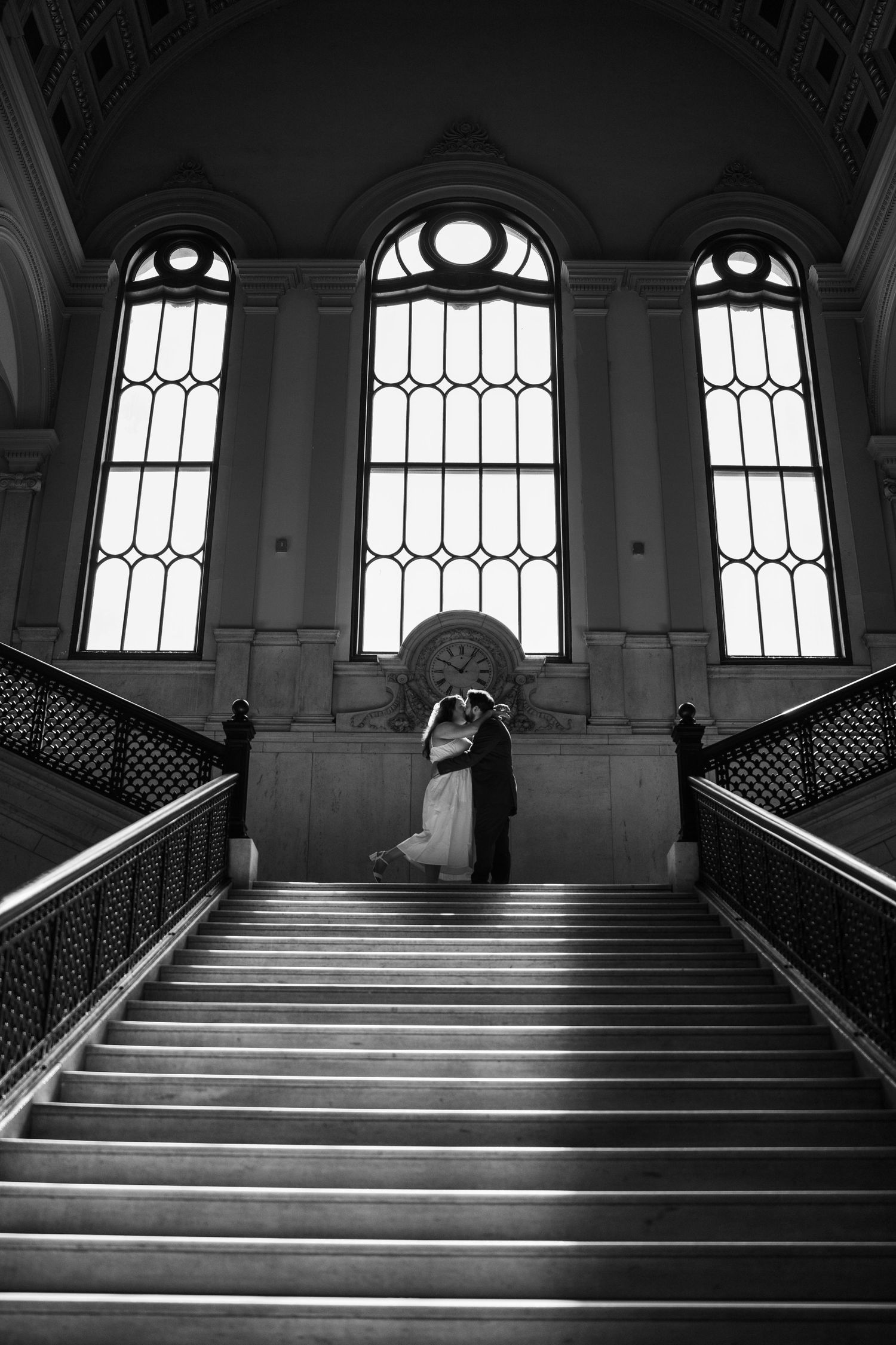 Black and white photo of a bride and groom embracing on grand staircase with arched windows in elegant venue, Worcester City Hall.