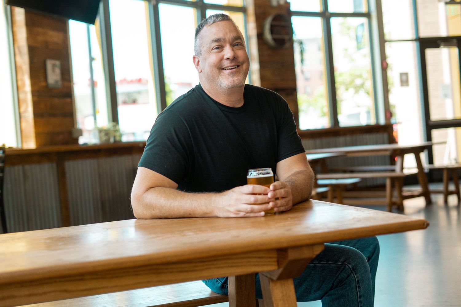 Smiling man in black t-shirt holding a craft beer glass at a wooden table in a modern brewery restaurant.