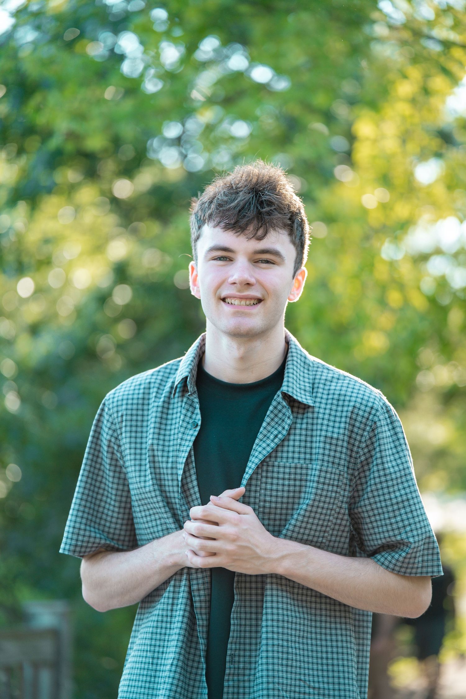 Smiling young man in a green plaid shirt standing outdoors with lush green trees in the background.