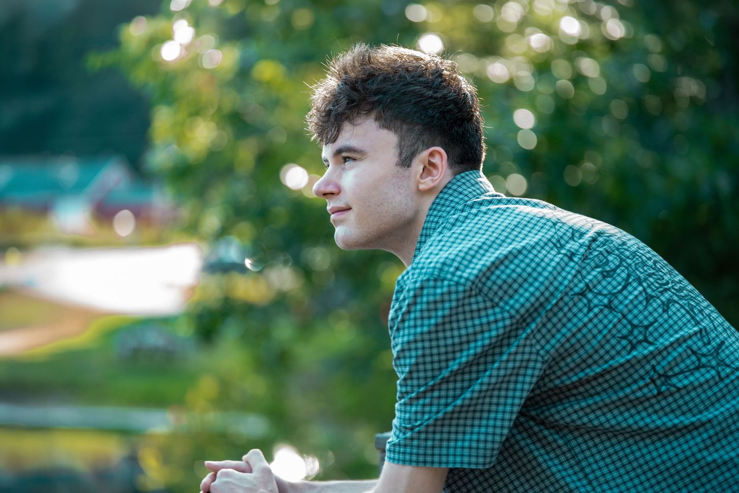 Young man in green plaid shirt gazing thoughtfully outdoors with lush green trees in background.