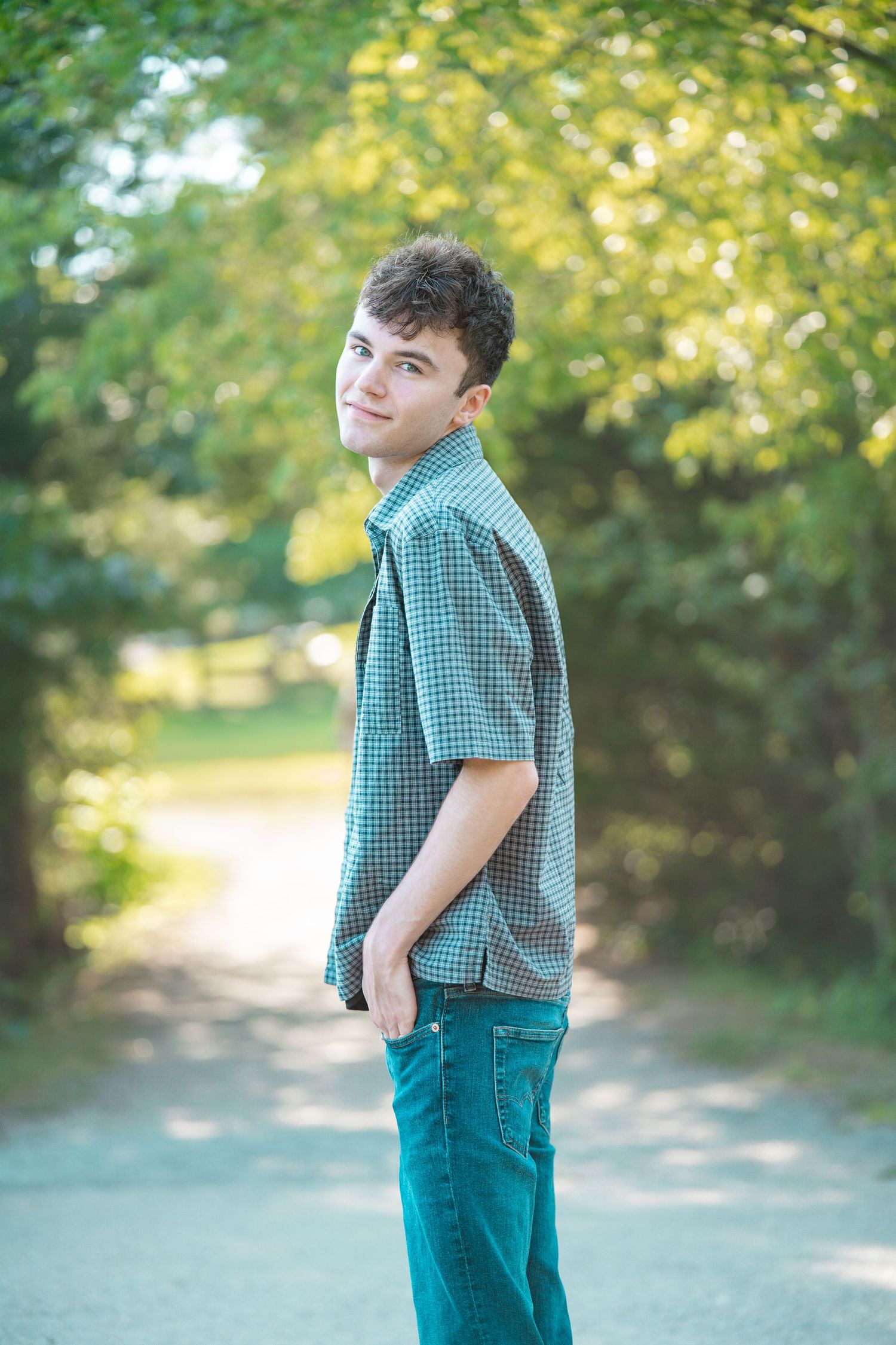 Young man in green plaid shirt and jeans standing on a tree-lined path, smiling over his shoulder. Blackstone Heritage and Canal State  Park