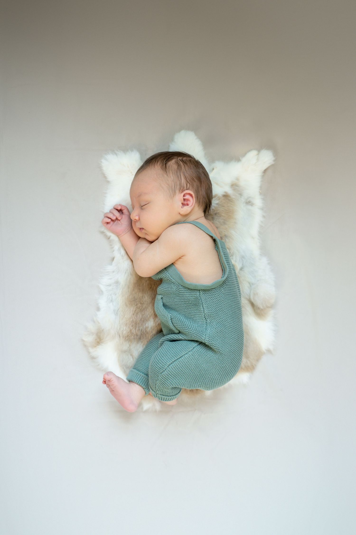 Newborn baby sleeping peacefully on white fur rug wearing a sage green romper, photographed from above.