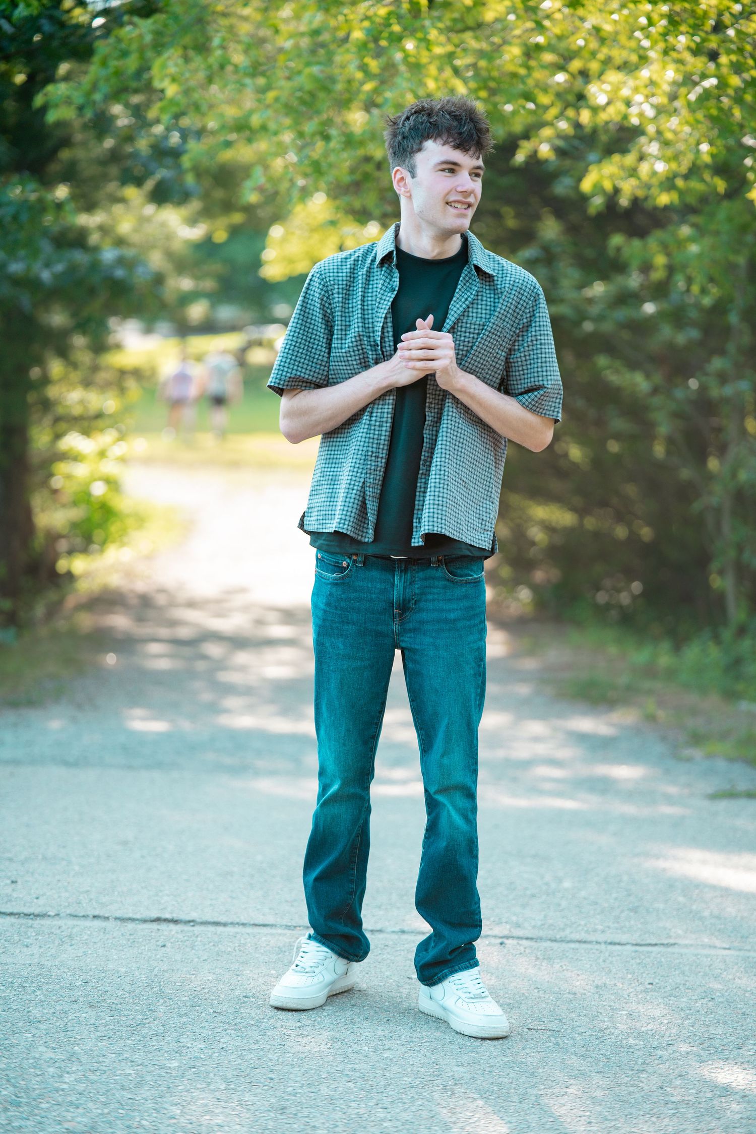 Young man in green plaid shirt and jeans smiling on a tree-lined path outdoors.