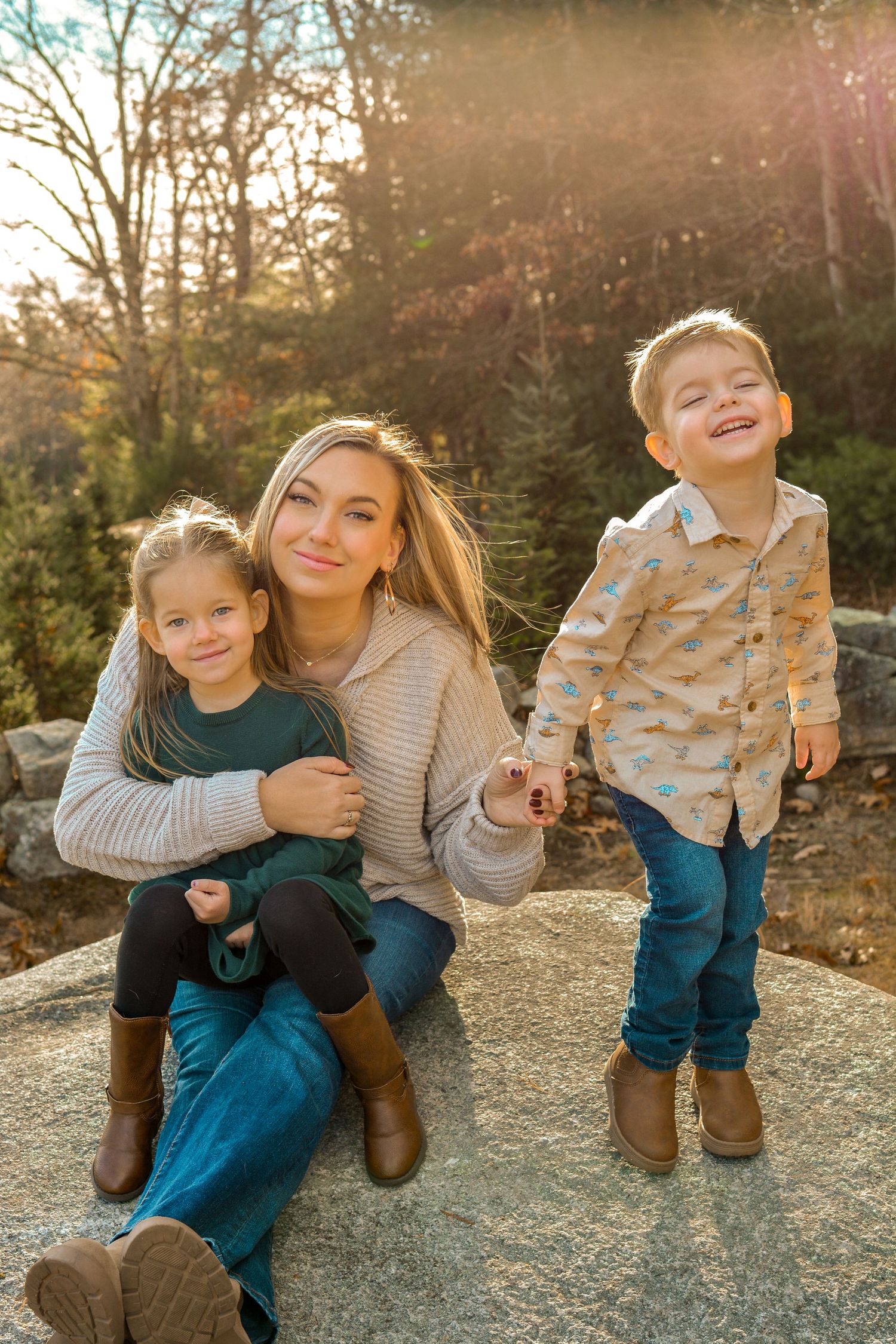 A woman sits on a rock outdoors holding two young children, smiling warmly in golden autumn sunlight.