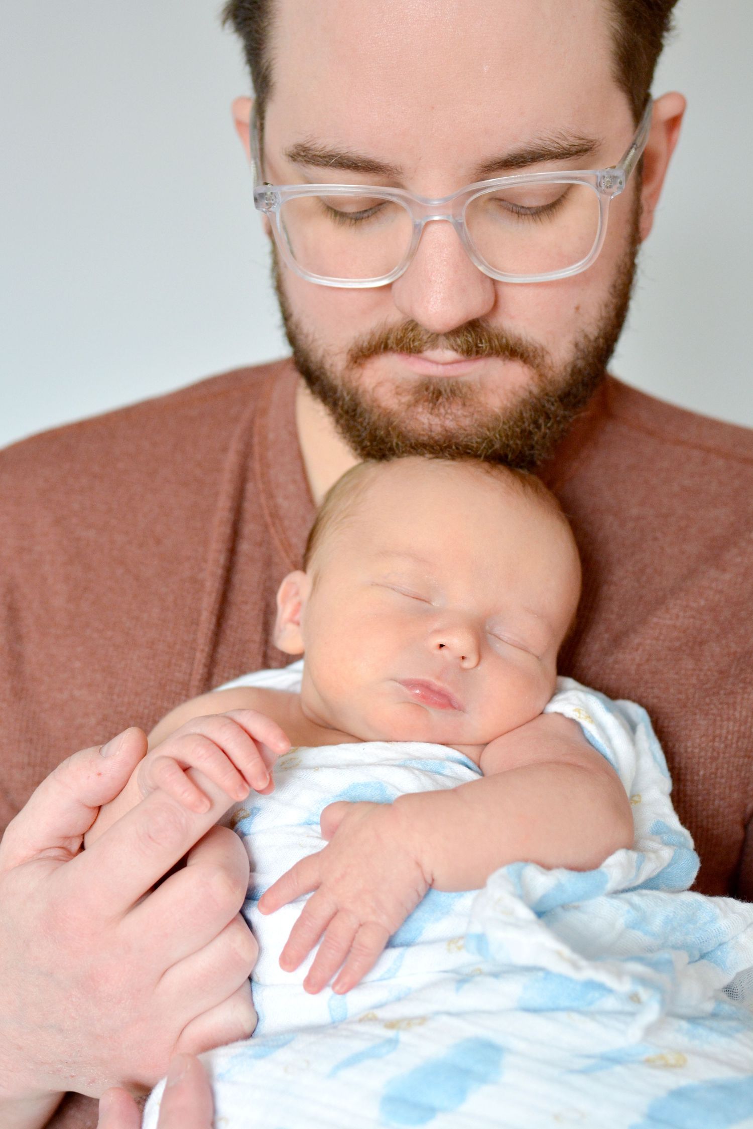 Bearded man with glasses tenderly holding a sleeping newborn baby wrapped in a blue blanket.