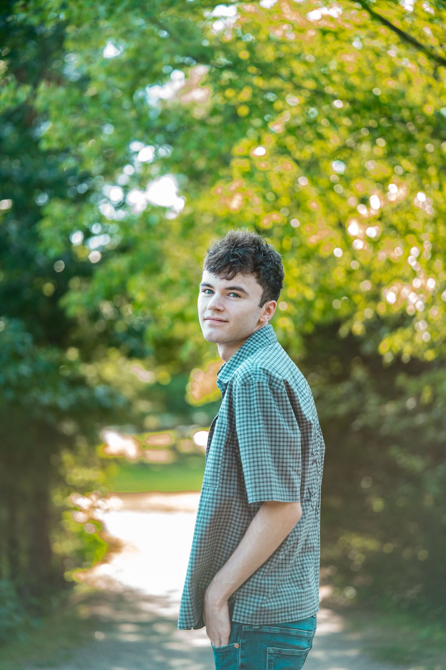 Young man in a plaid shirt standing on a tree-lined path, looking back over his shoulder on a sunny day.