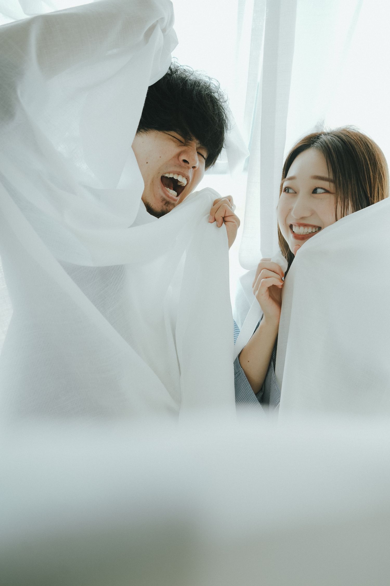 カーテン越しに笑顔をのぞかせるご夫婦の自然なポートレート