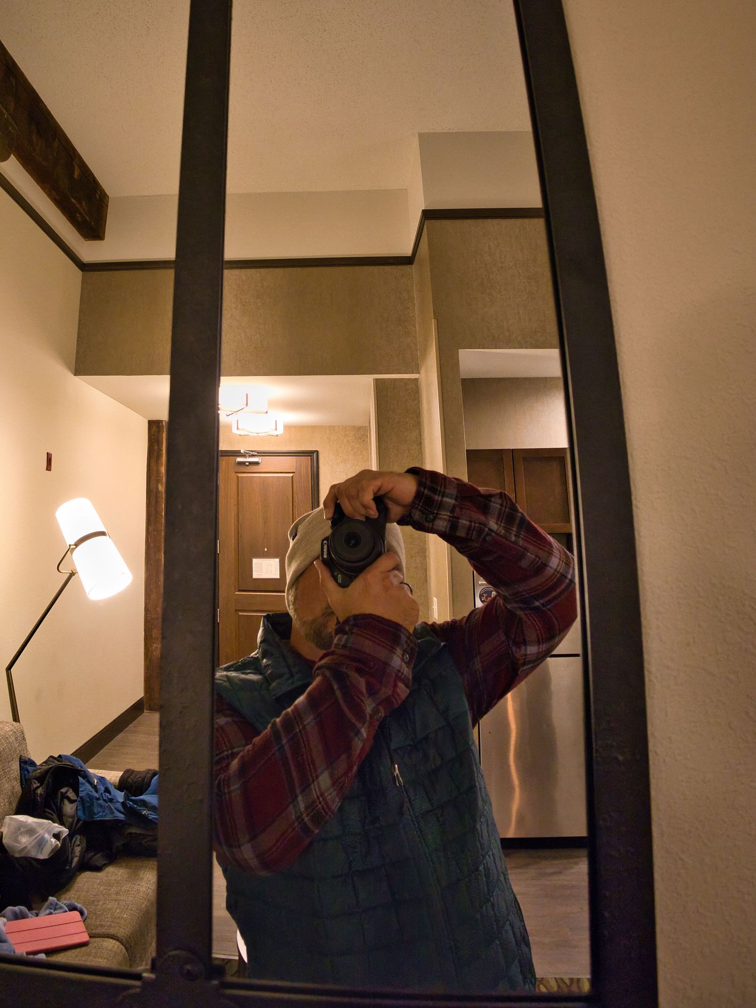 Photographer taking a mirror selfie with a camera in a warmly lit hotel room wearing a plaid shirt and blue vest.