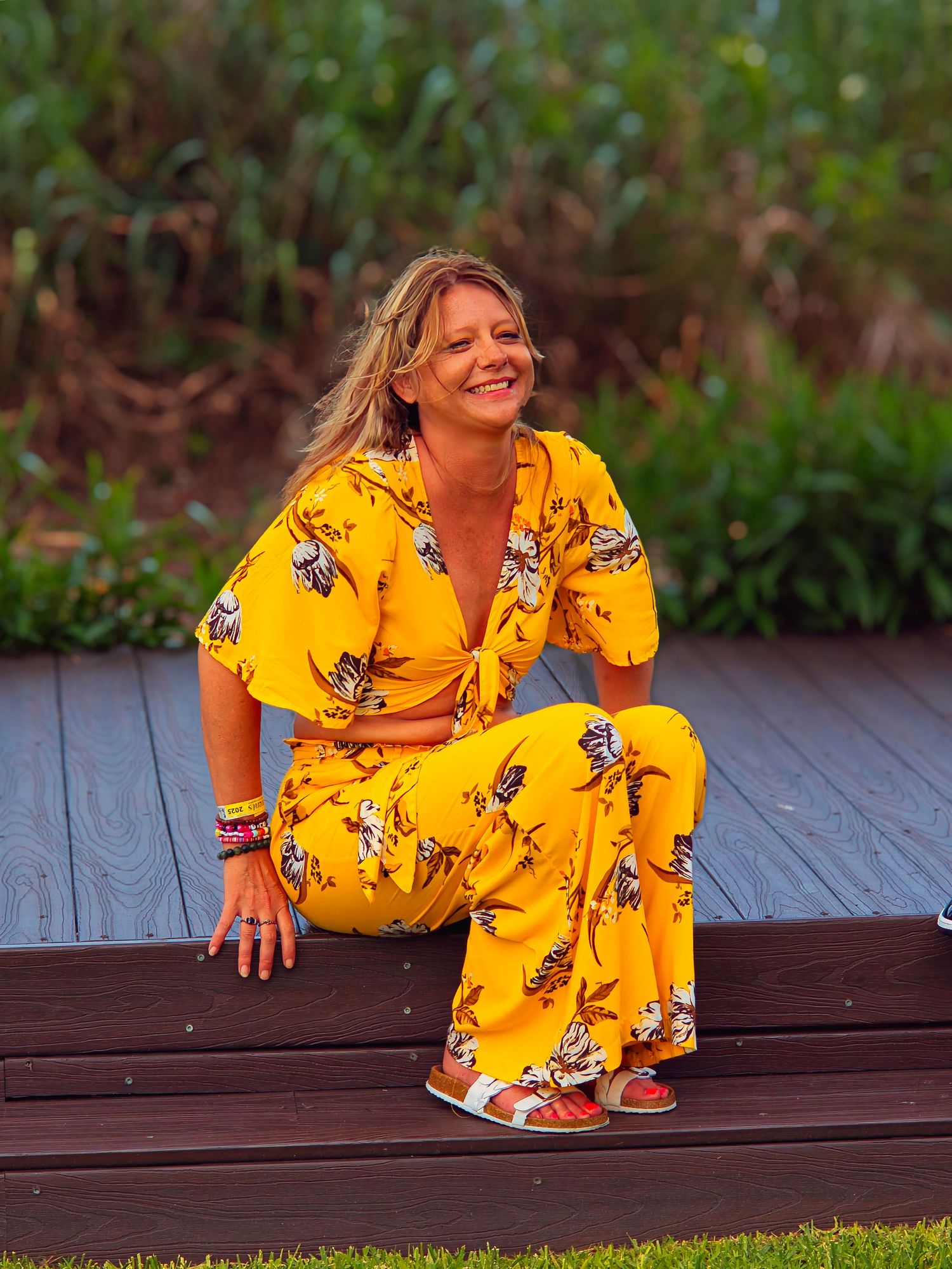 Cheerful blonde woman in yellow floral outfit sitting on wooden deck outdoors smiling.