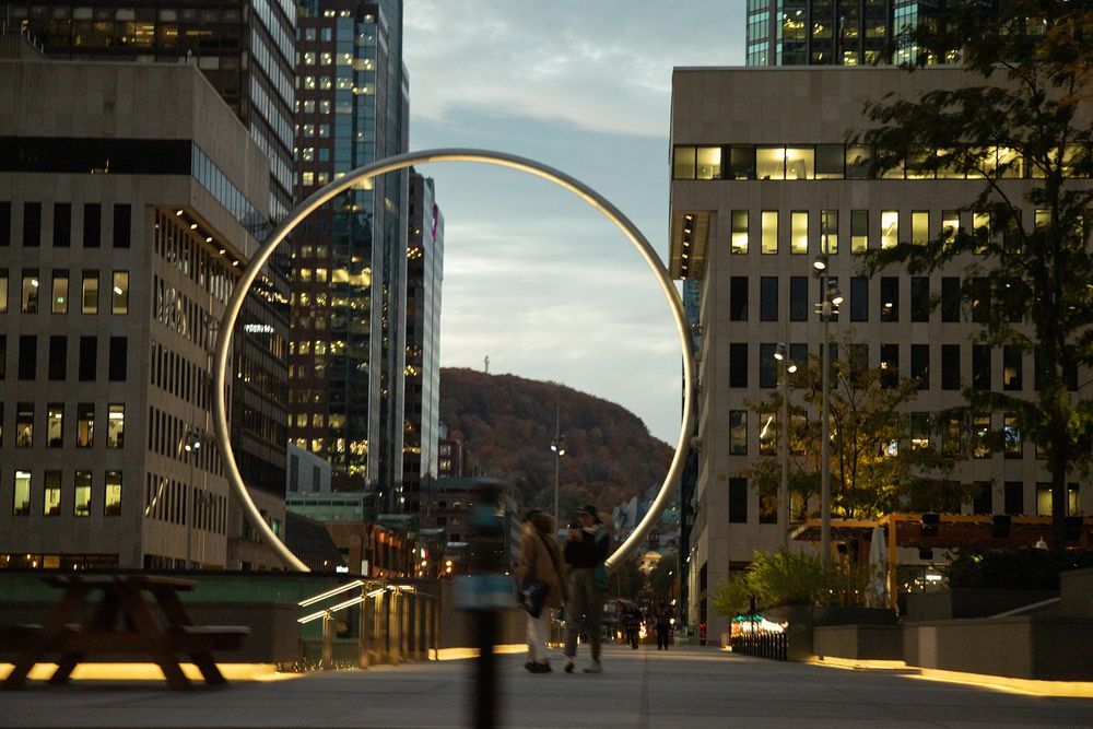 Large illuminated circular art installation on a city street at dusk, with glowing skyscrapers and a mountain backdrop.
