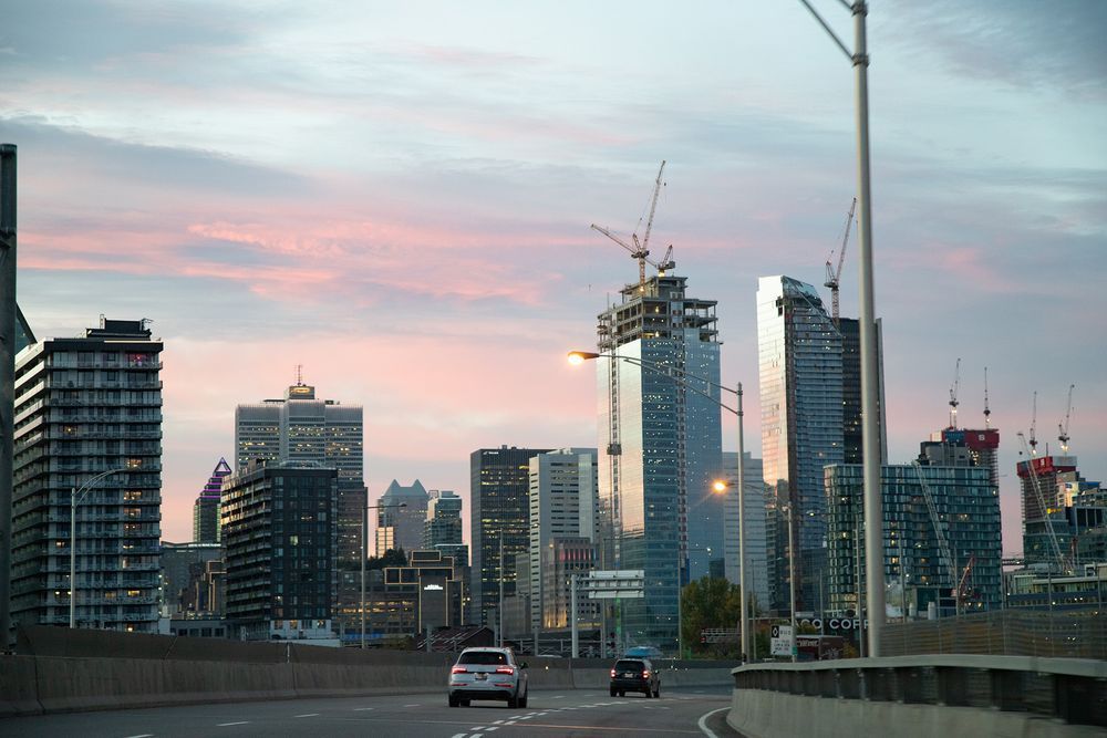 City skyline at dusk with cranes, skyscrapers, and cars on a highway under a pink and blue sky.