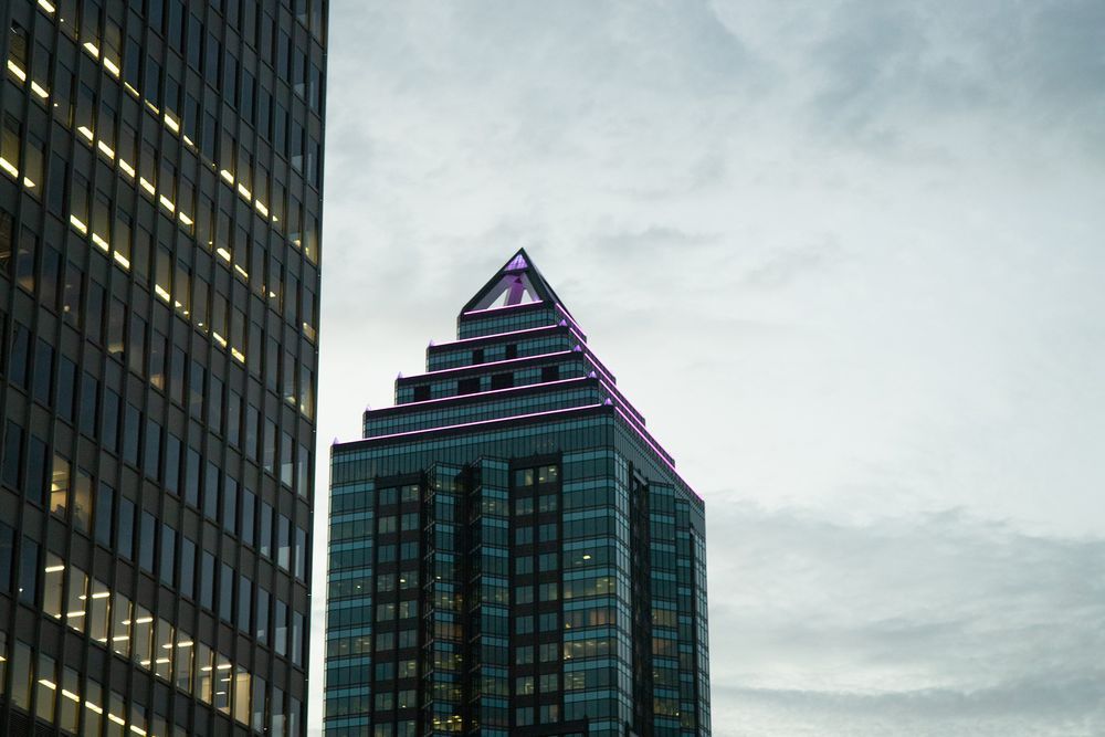 Modern skyscrapers with a pyramid-topped glass tower and illuminated office building against a cloudy sky.