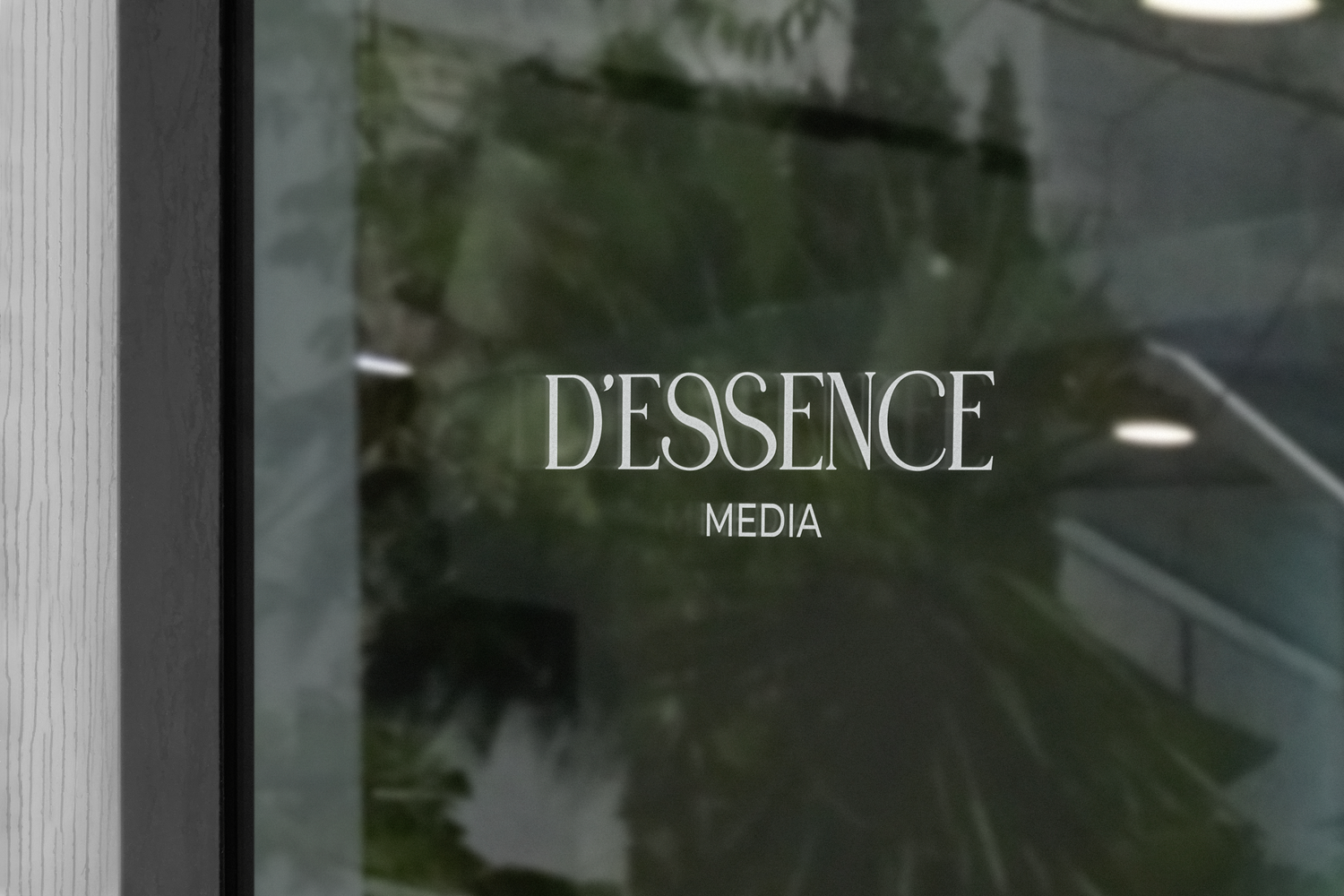 D Essence Media logo etched on glass window with green foliage reflection.