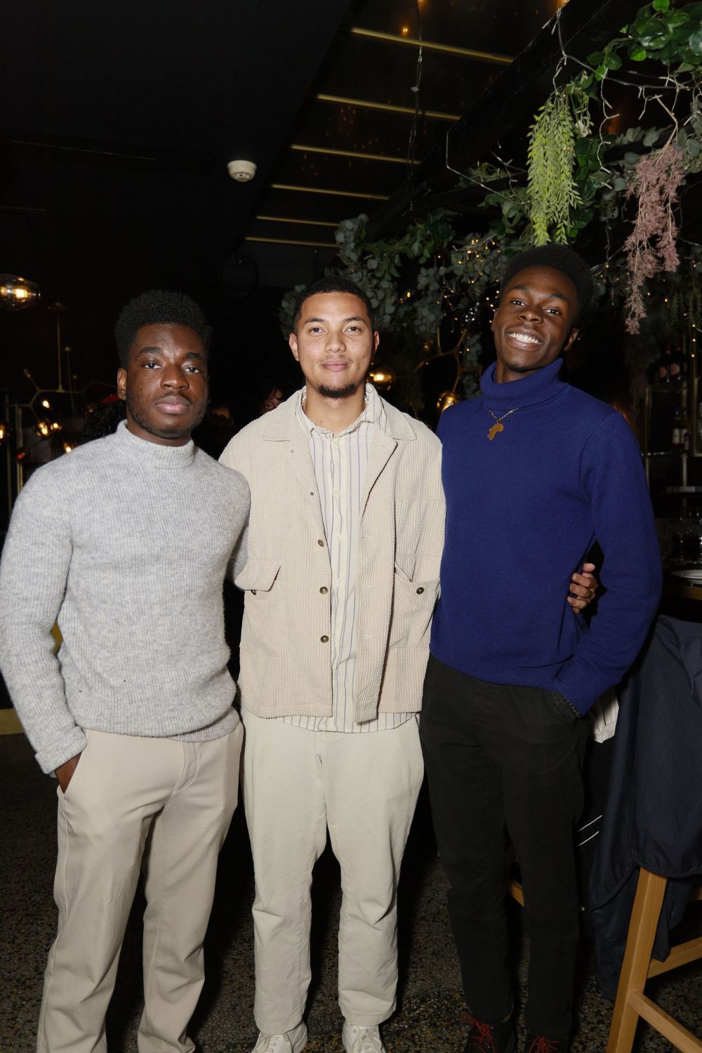 Three young men smiling together at an indoor evening social event surrounded by greenery decor.