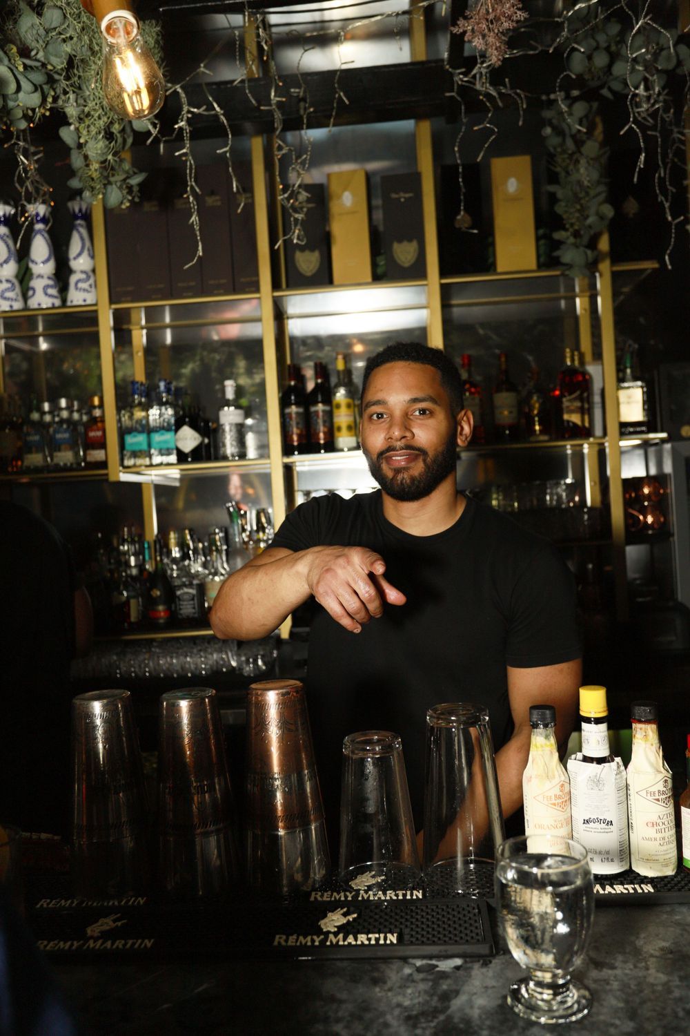 Bartender in black shirt posing behind bar with Remy Martin bottles and glassware.
