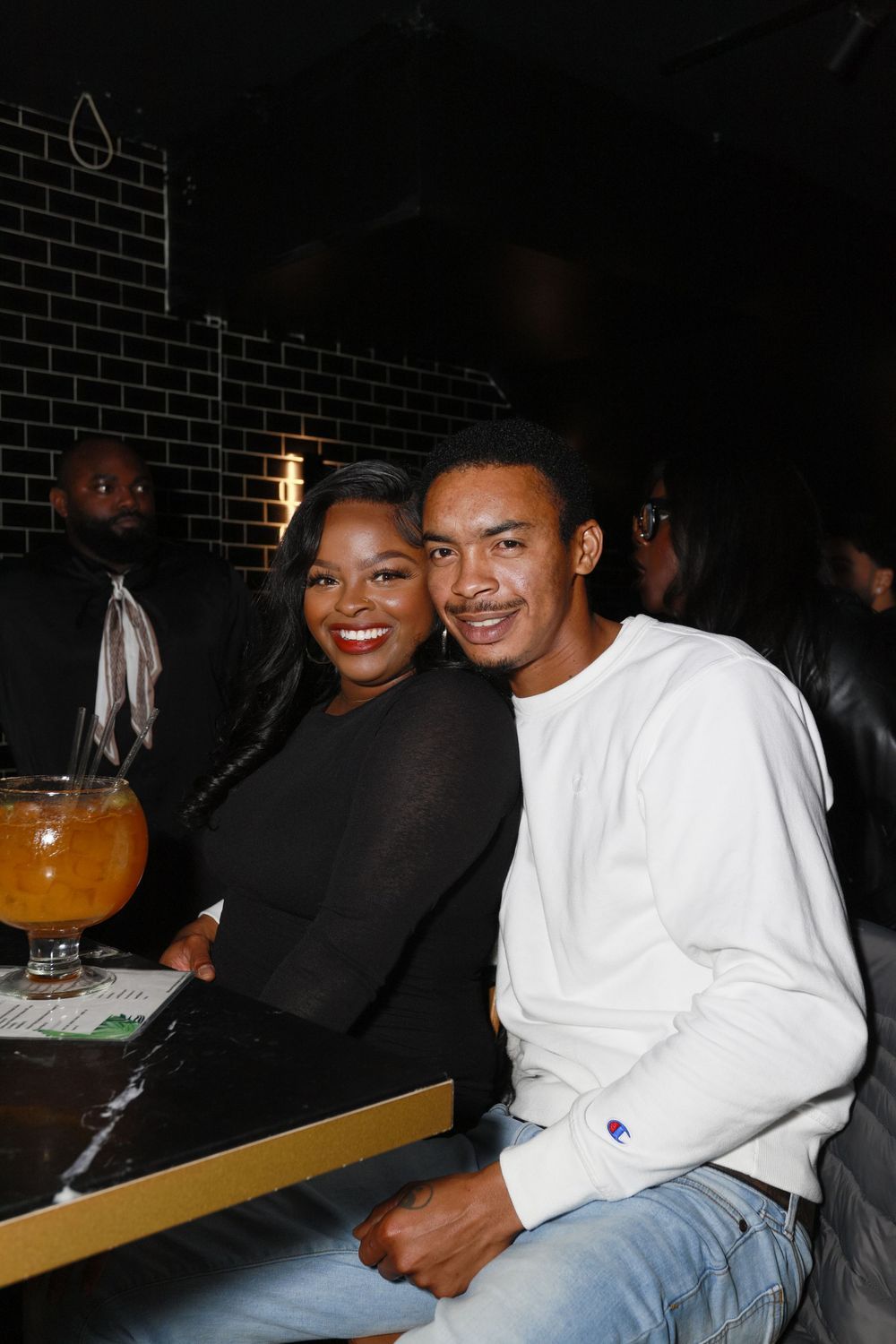 Couple smiling together at a dark bar with orange cocktail drinks nearby.