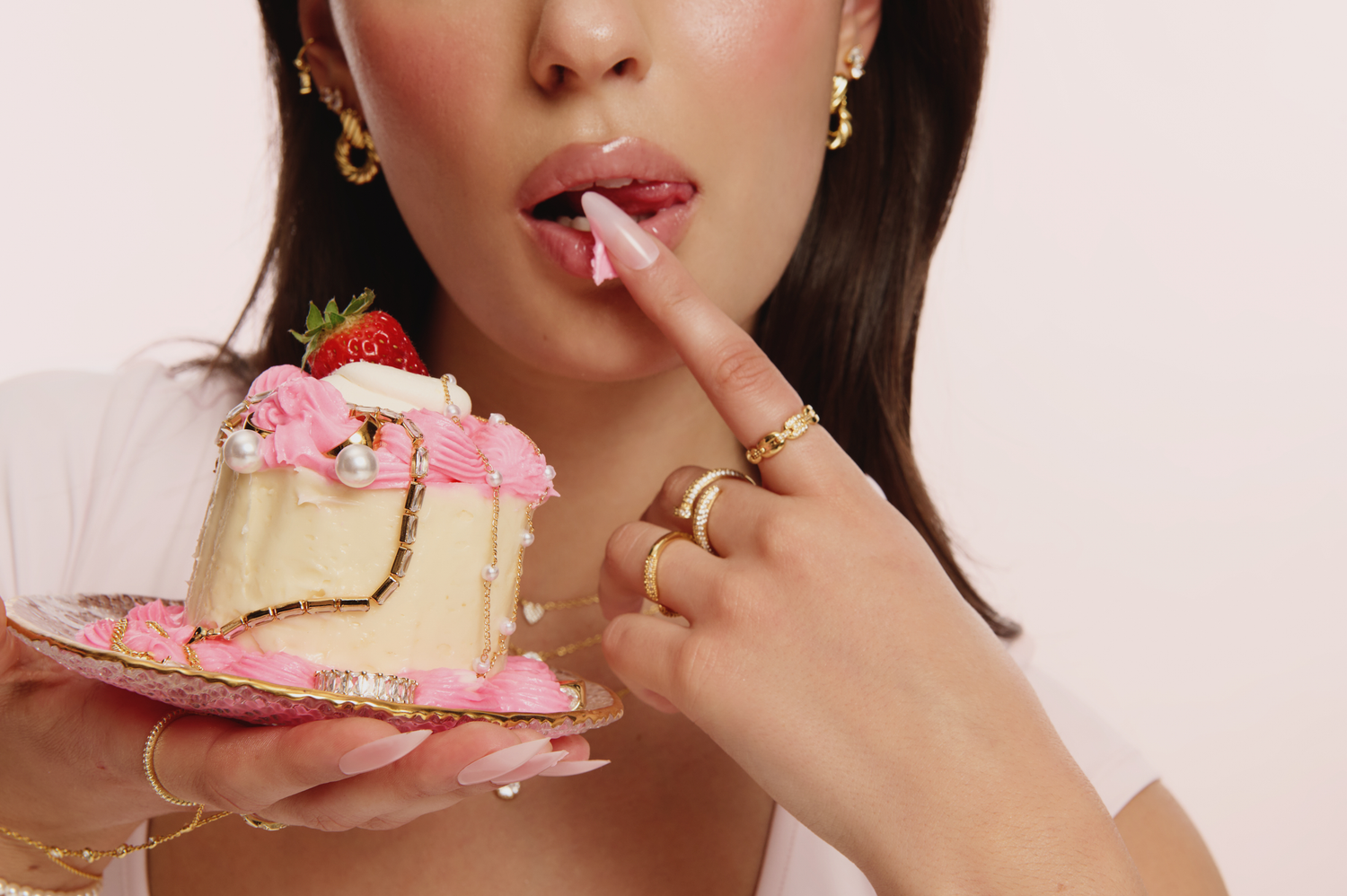 Woman holding a pink strawberry cake slice while tasting frosting off her finger, wearing gold rings and earrings.
