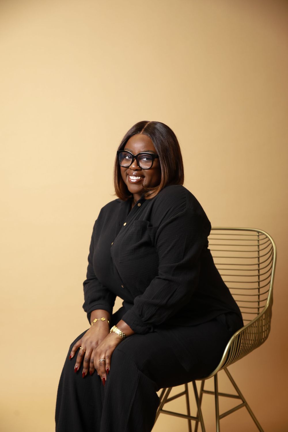 Smiling woman in black outfit seated on gold wire chair against beige background.