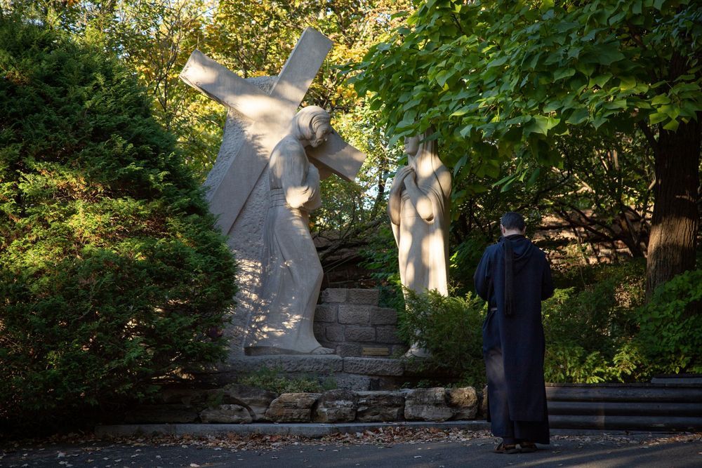 A robed figure prays before a stone cross-bearing statue surrounded by lush green trees in a serene garden setting.
