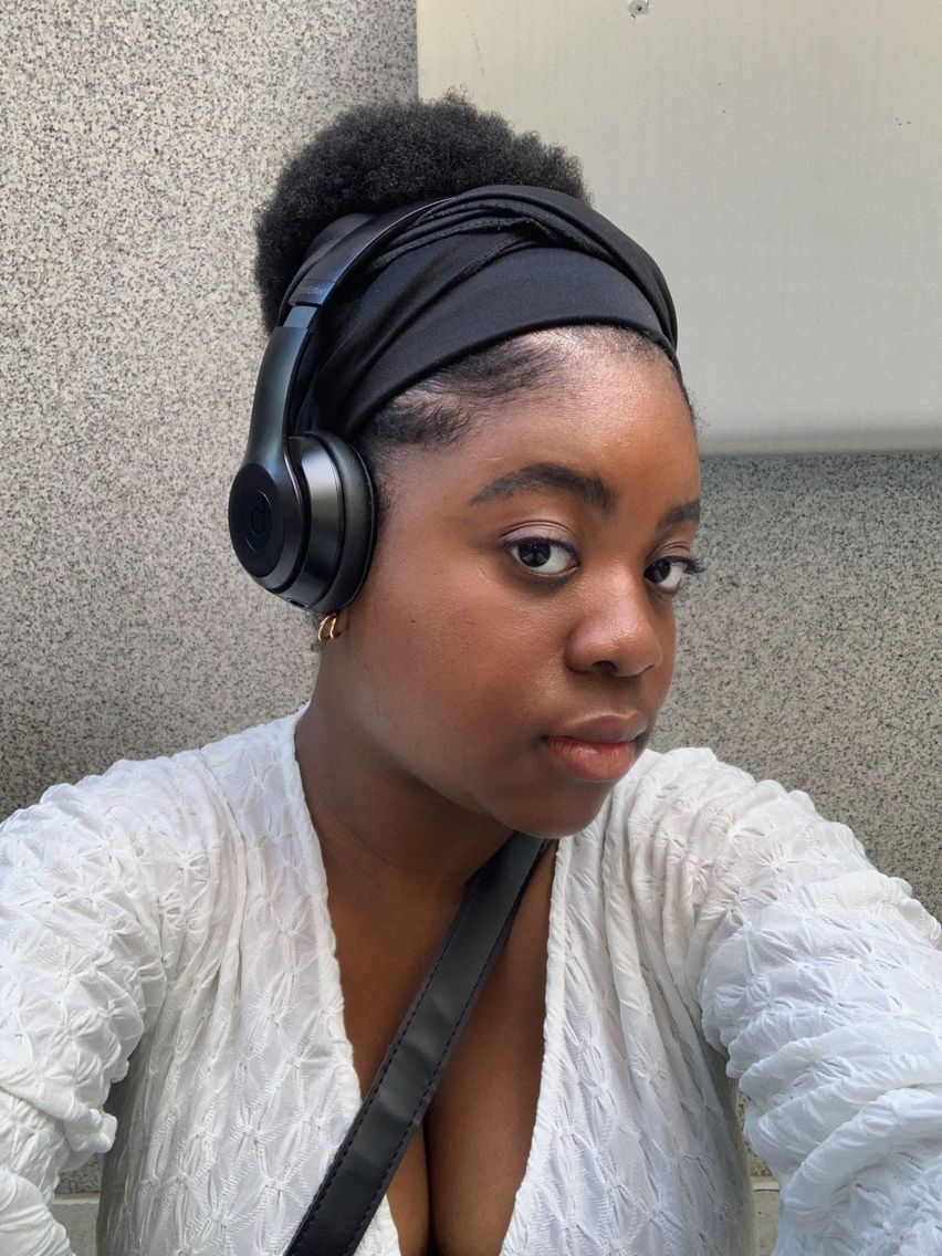 Young woman wearing black over-ear headphones and a black headband, dressed in a white textured top, taking a selfie.