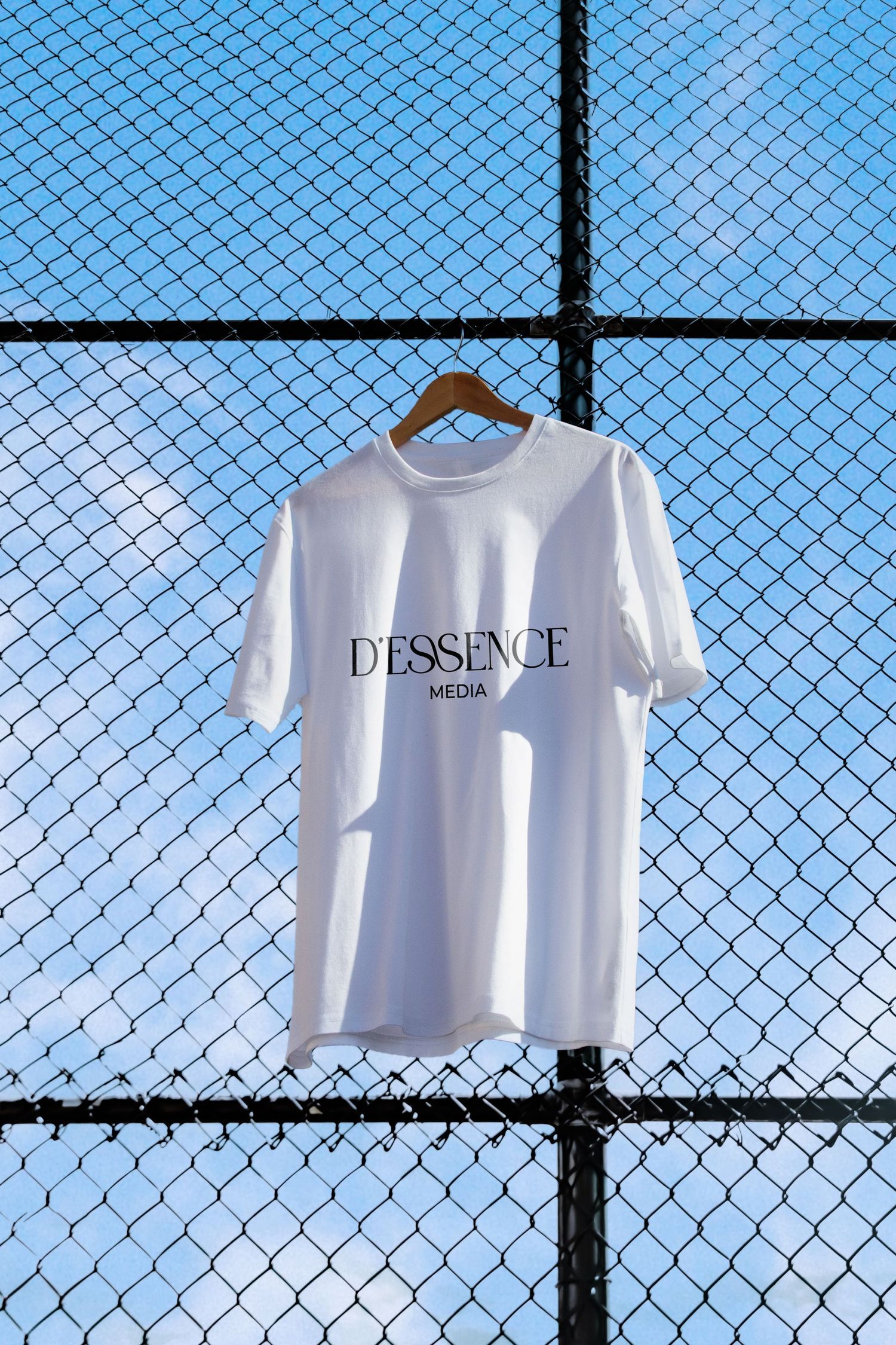 White Dessence Media t-shirt hanging on wooden hanger against blue sky chain-link fence.