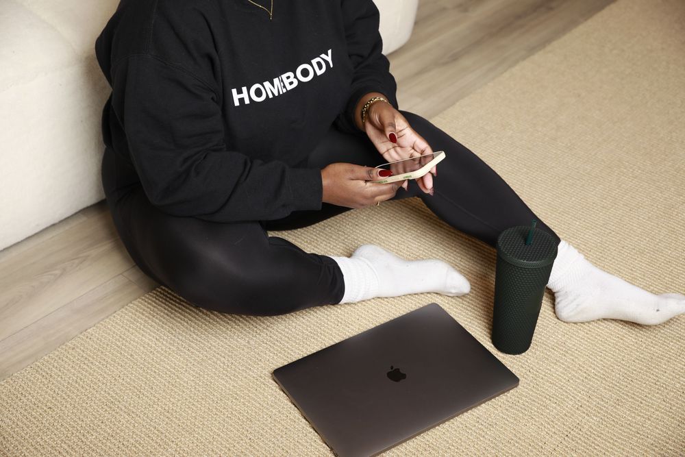 Person in black Homebody sweatshirt sitting cross-legged on floor with laptop and tumbler.