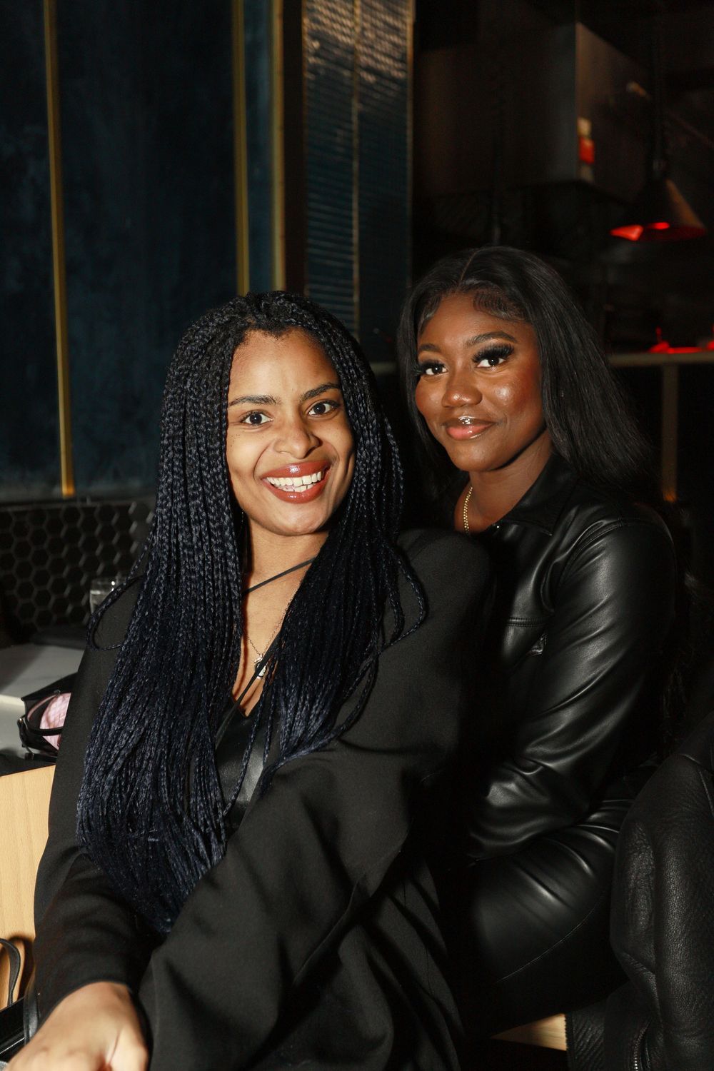 Two women smiling together at a dimly lit social event, one in braids and black suit, one in leather jacket.